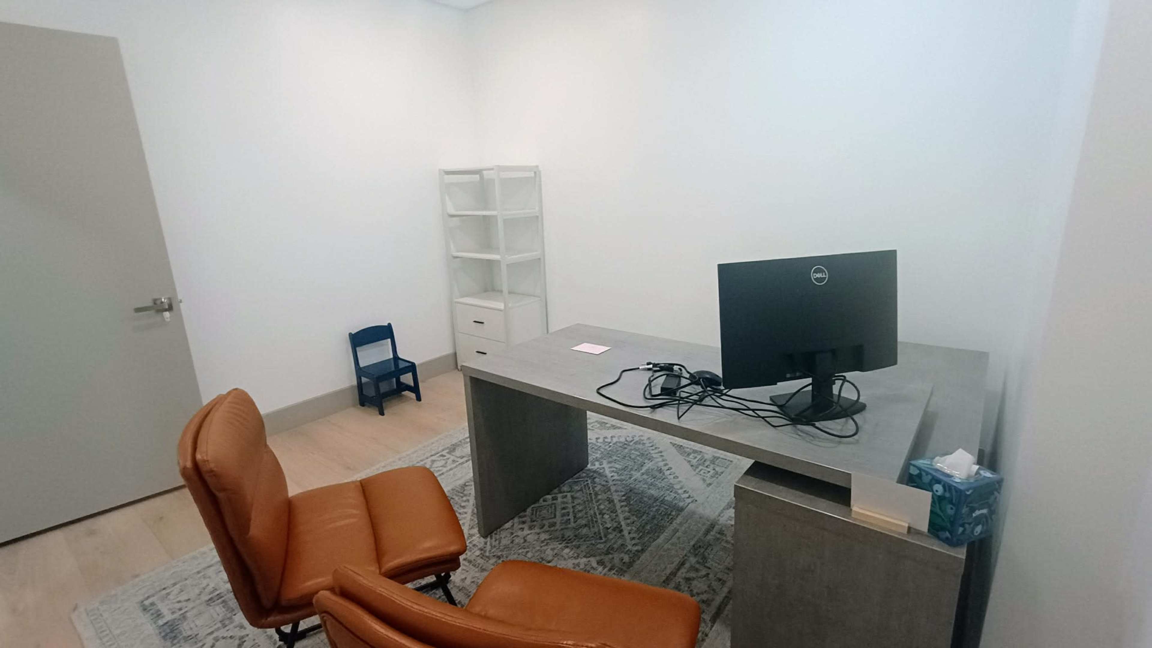 The image shows a small, minimalist office with a desk, a computer monitor, two brown chairs, and a white storage unit in the corner.
