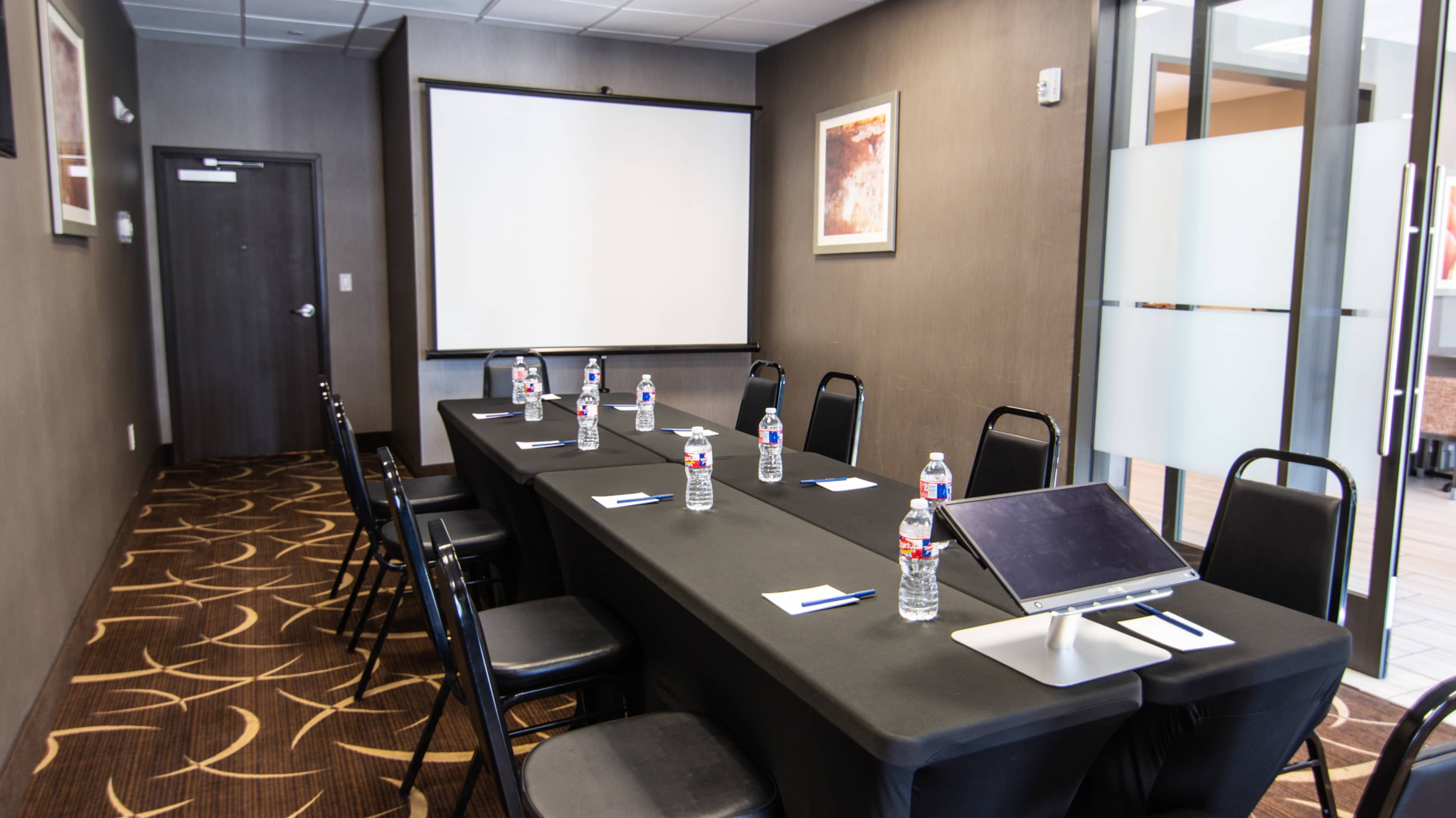 A conference room features a long table set with water bottles, notepads, and a monitor, with a blank projection screen at one end.