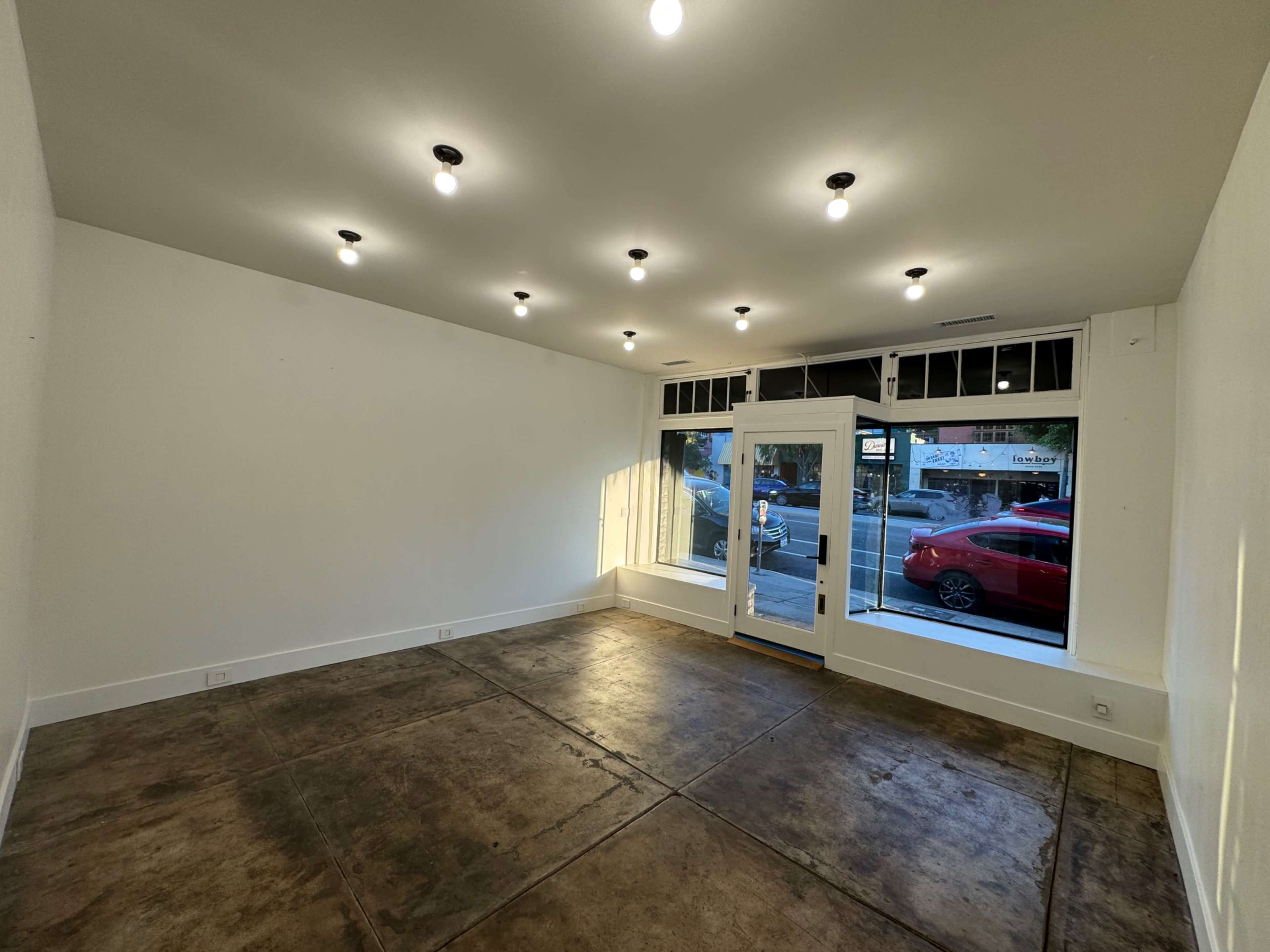 An empty room with a concrete floor and bright lighting, featuring a large front window and a glass door leading outside.