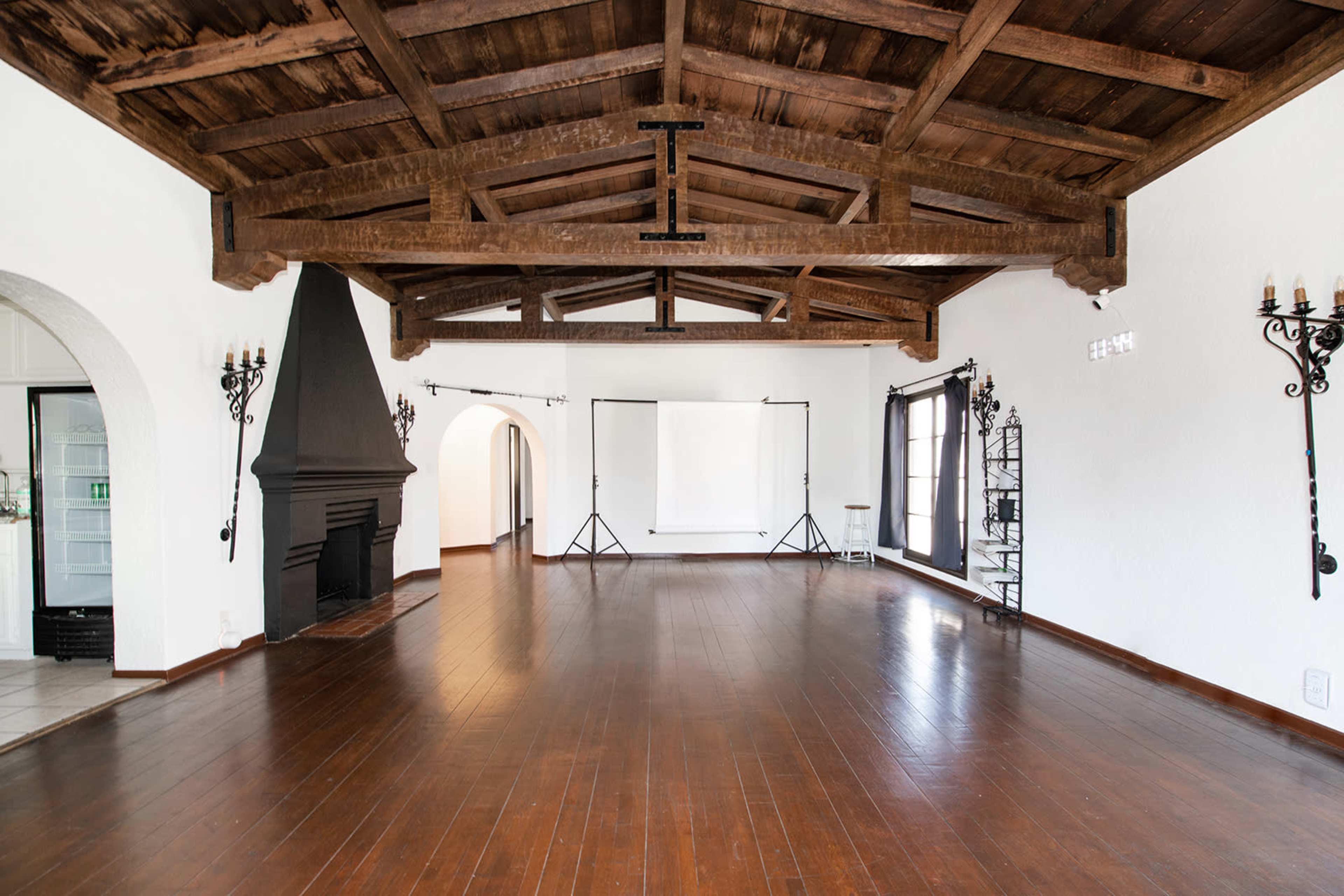 A spacious room with wooden beams on the ceiling, a fireplace, and large windows, featuring polished wooden floors and basic lighting equipment set up in the background.