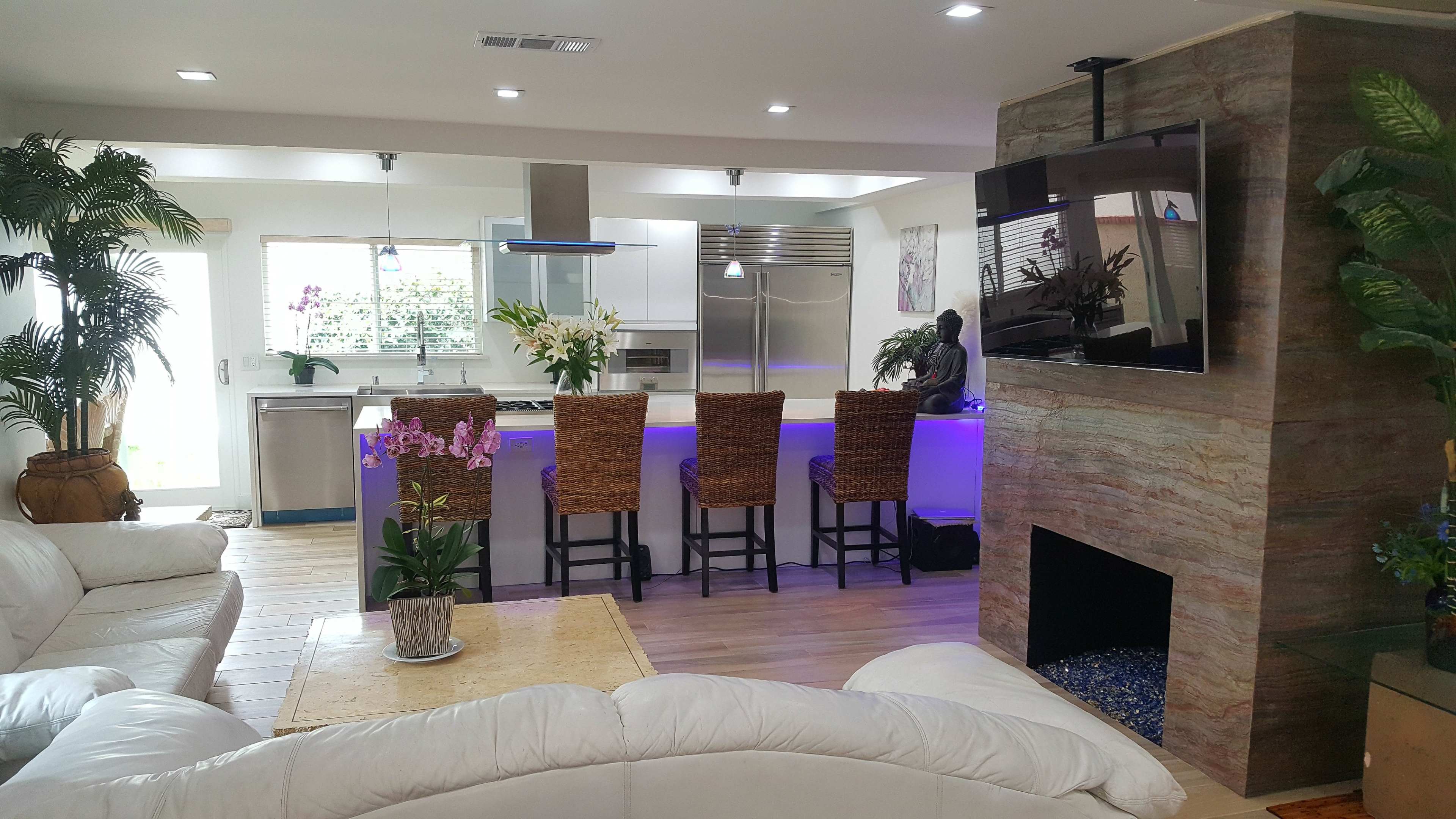 A modern living room with a white sectional sofa, a coffee table, and a kitchen area featuring bar stools, a large island, and a wall-mounted television above a stone fireplace.