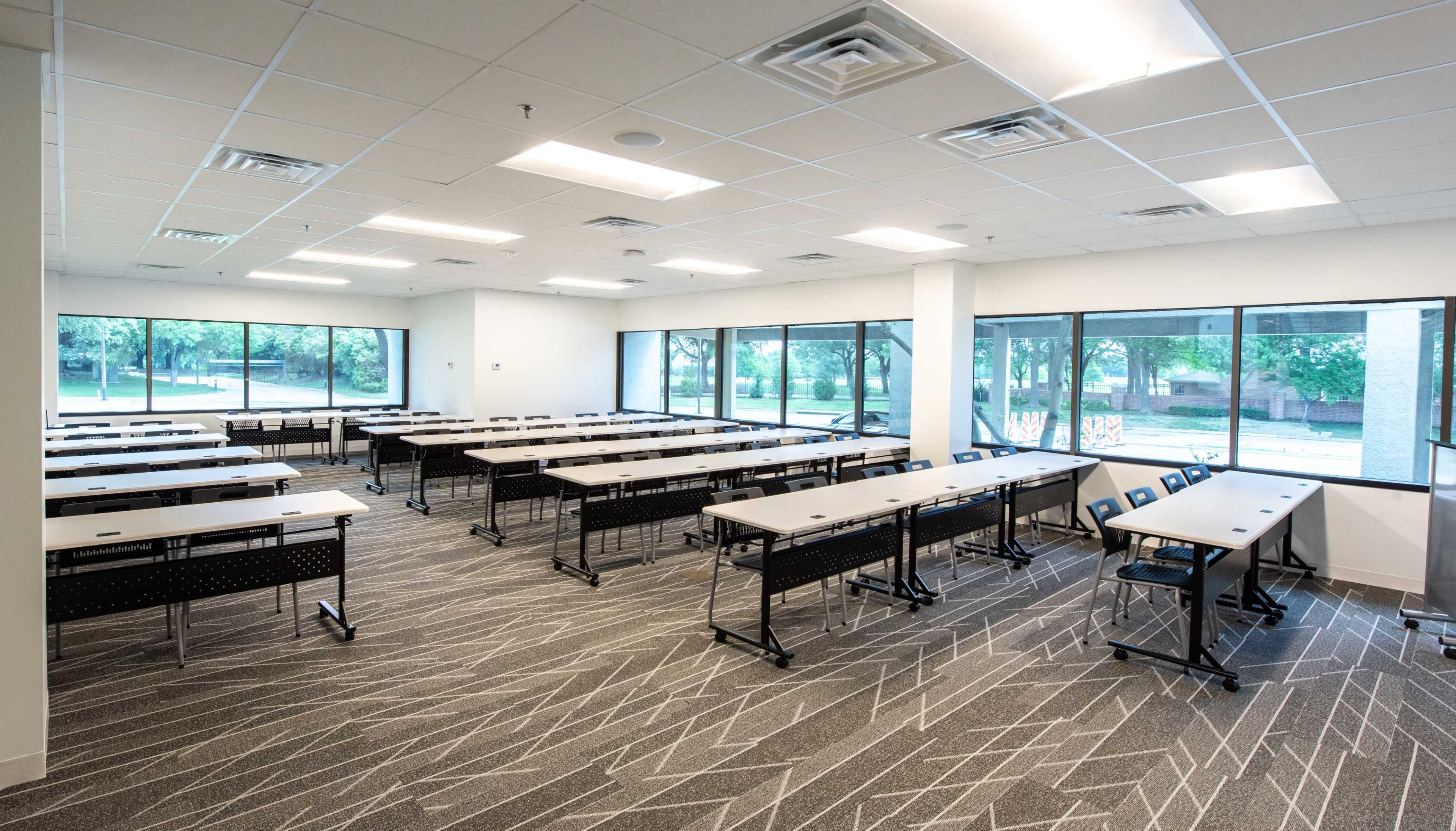 A spacious classroom arranged with several rows of tables and chairs, illuminated by large windows that provide natural light.