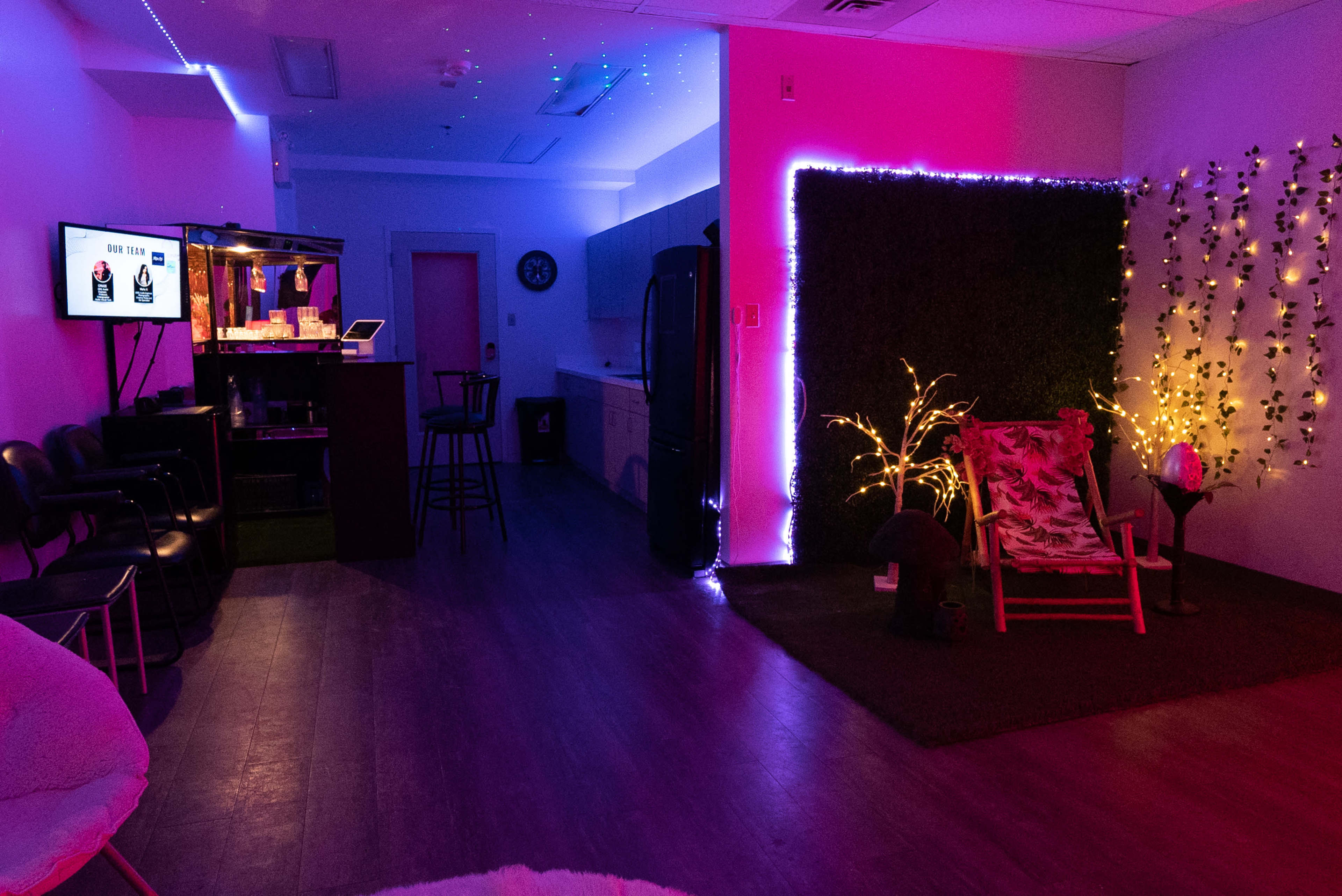 A lounge area with colorful lighting, a bar counter, a kitchen space, and a decorated wall featuring plants and fairy lights.
