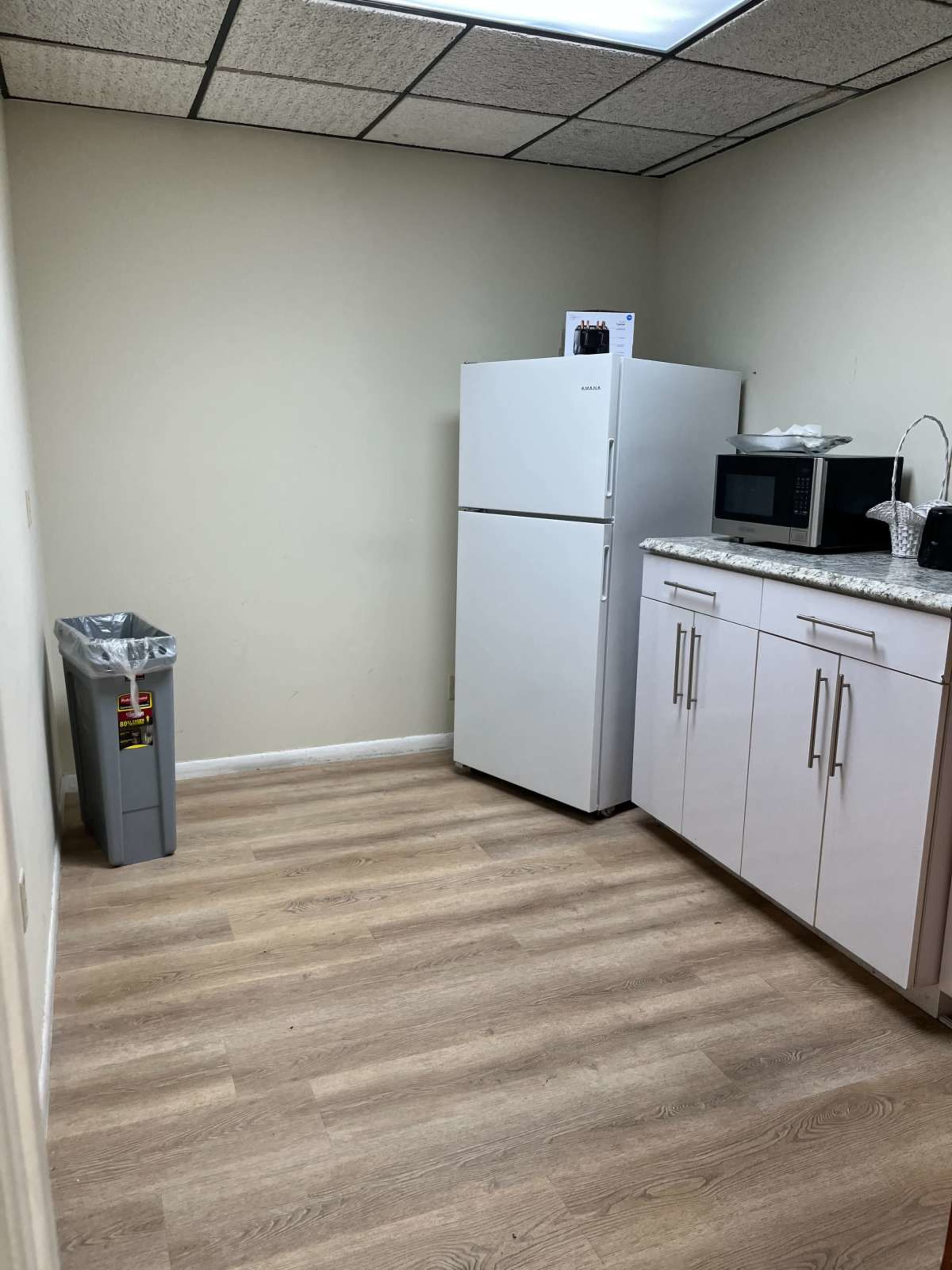 The image shows a small, empty kitchen area with a refrigerator, microwave, and a trash can.