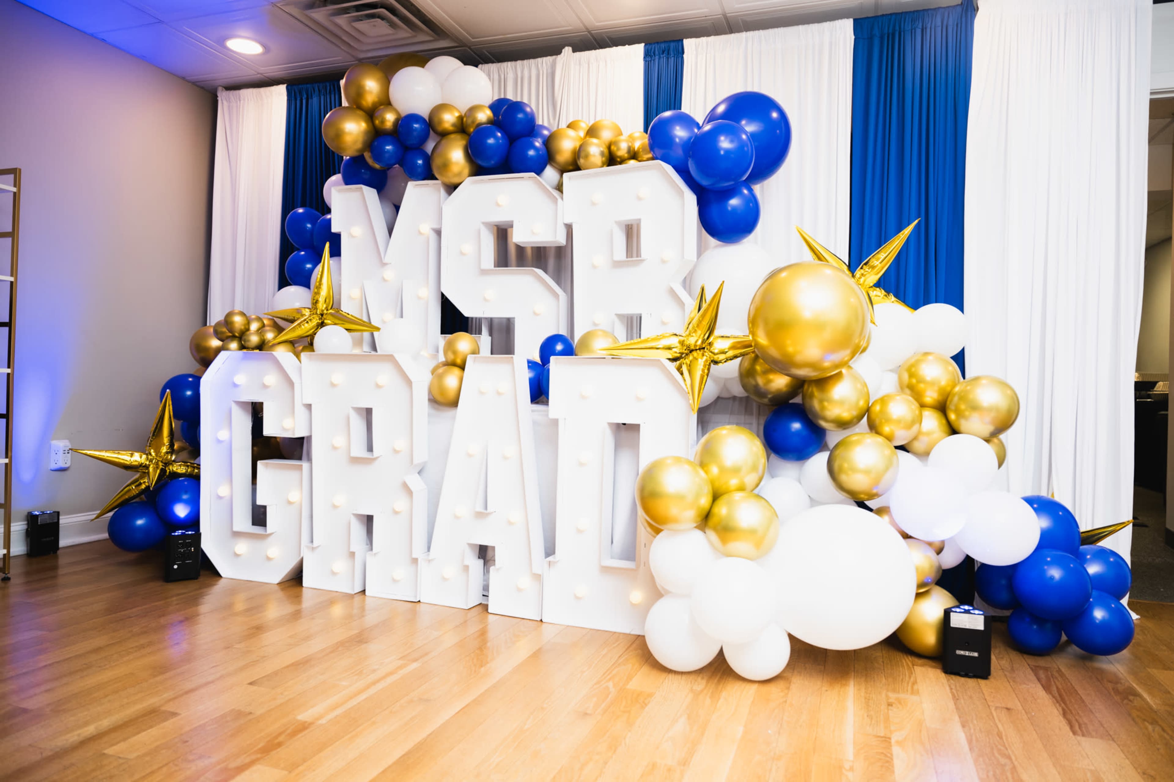 The image shows a decorative backdrop featuring large block letters spelling "MSB GRAD," surrounded by an arrangement of colorful balloons in blue, gold, and white.