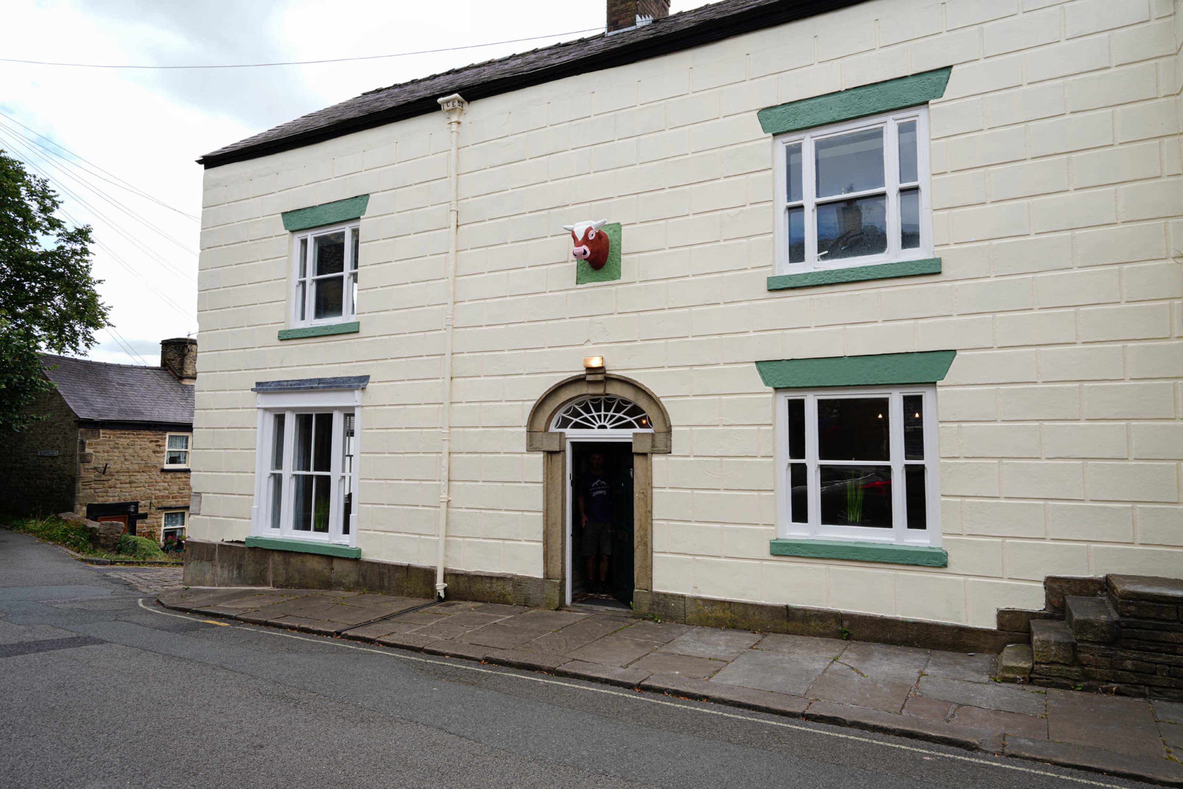 The image shows a two-story white building with green accents and a decorative head of a ram above the door, located at the corner of a street.