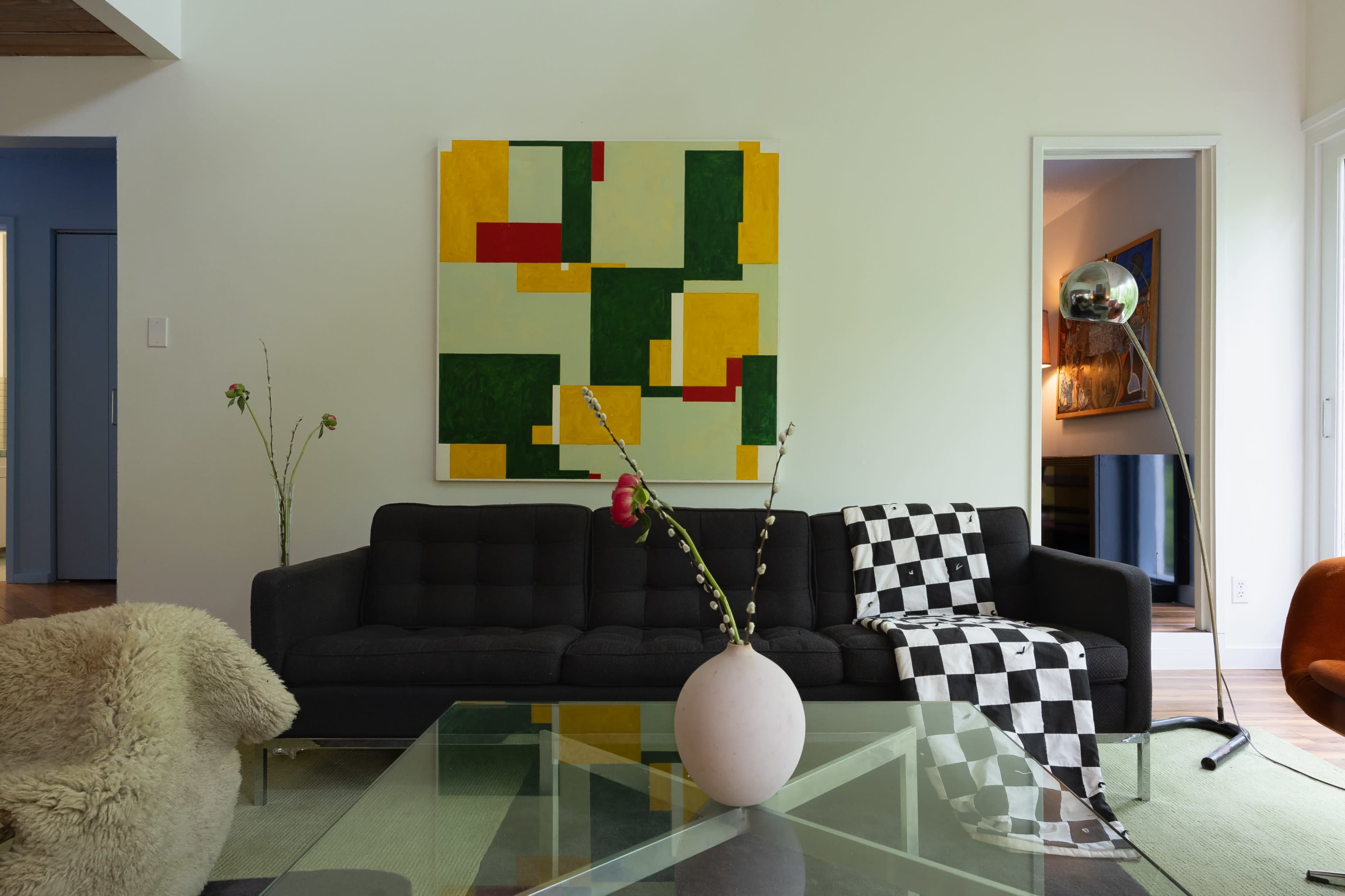 A modern living room features a black sofa, a geometric abstract painting on the wall, a round vase with flowers, and a glass coffee table.