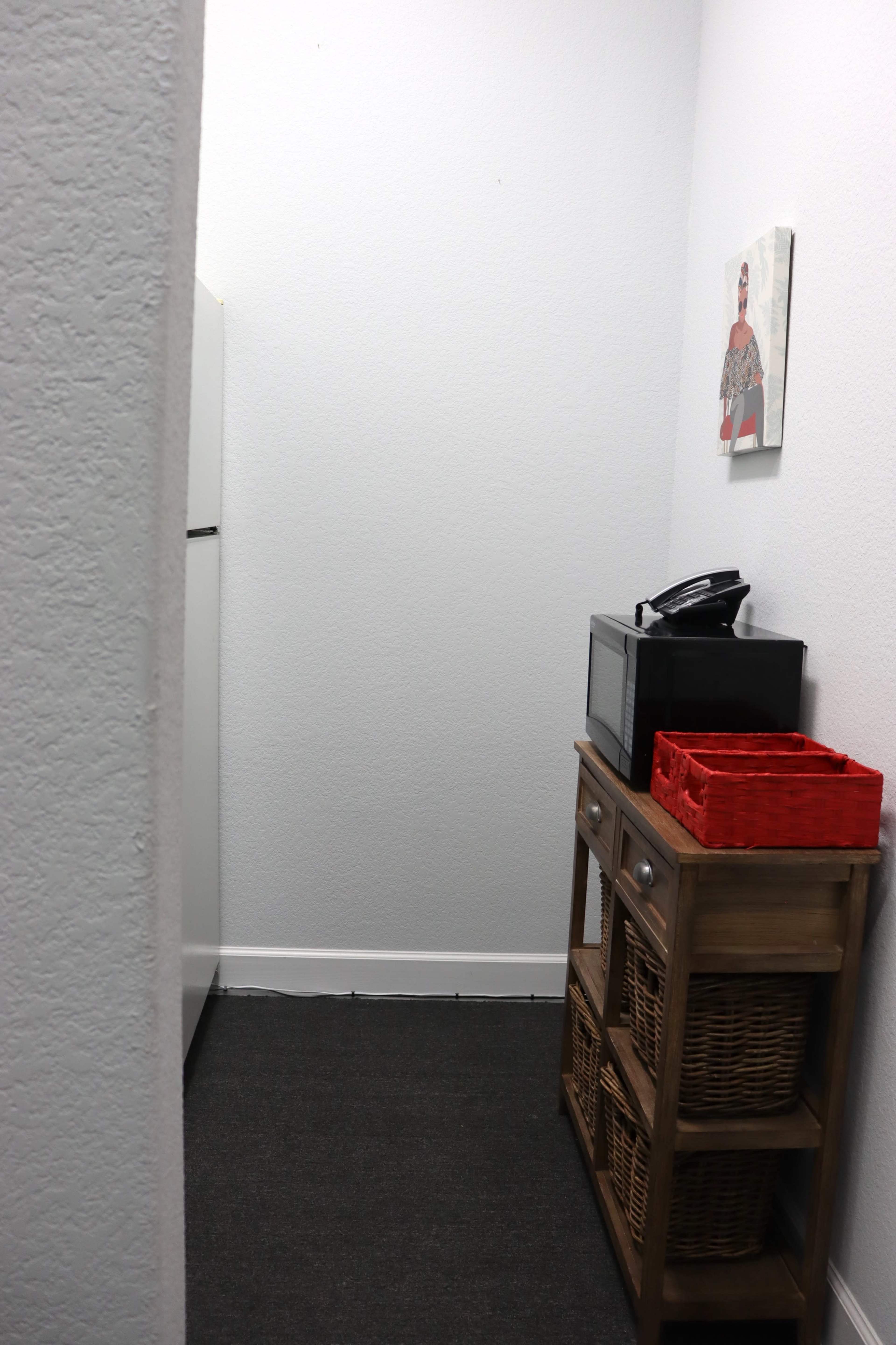 A narrow space features a refrigerator, a microwave on a wooden stand, and wicker baskets along with a framed picture on the wall.