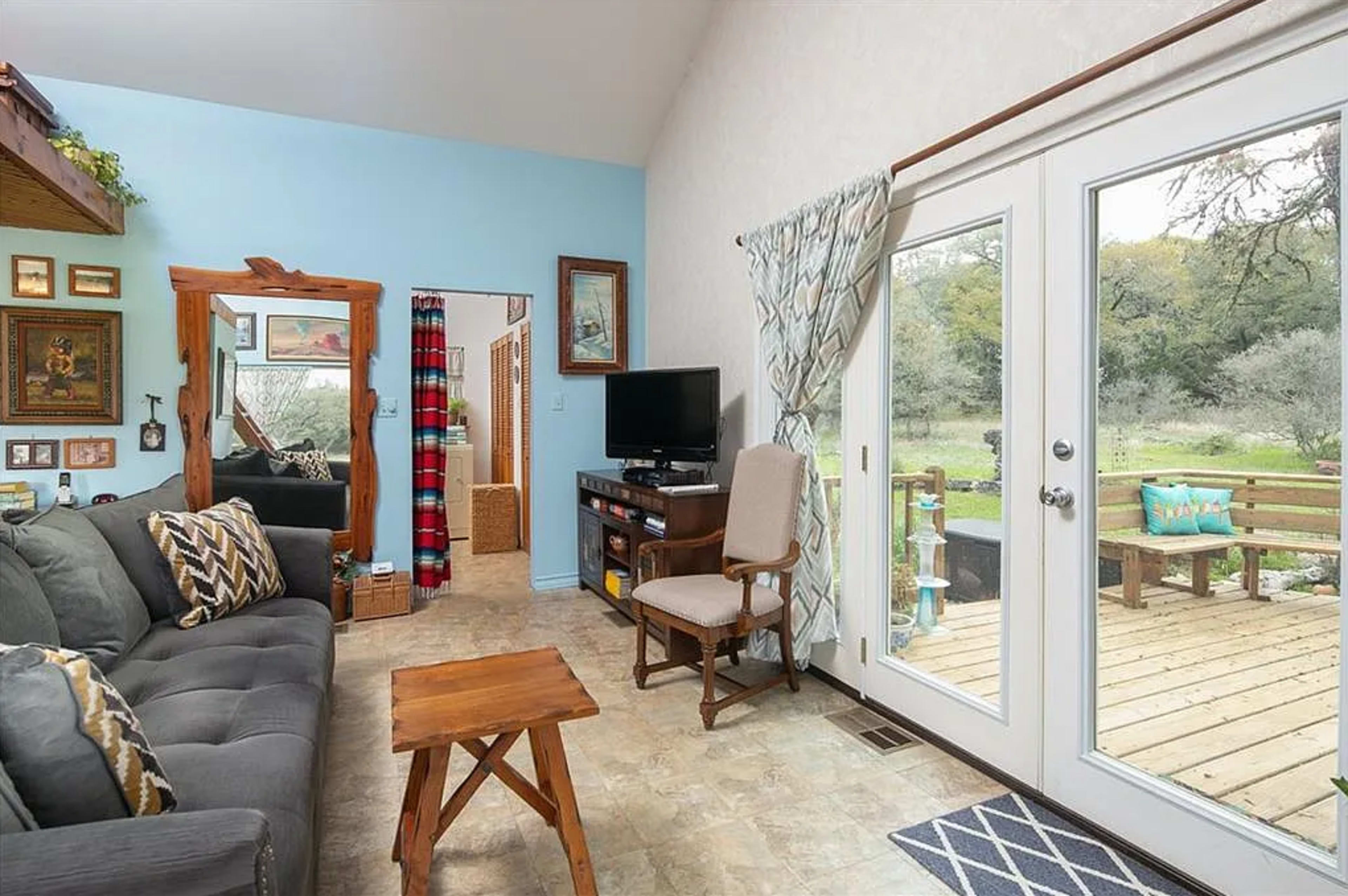 The image depicts a cozy living room with a gray couch, a small wooden table, and large glass doors opening to a wooden deck outside.