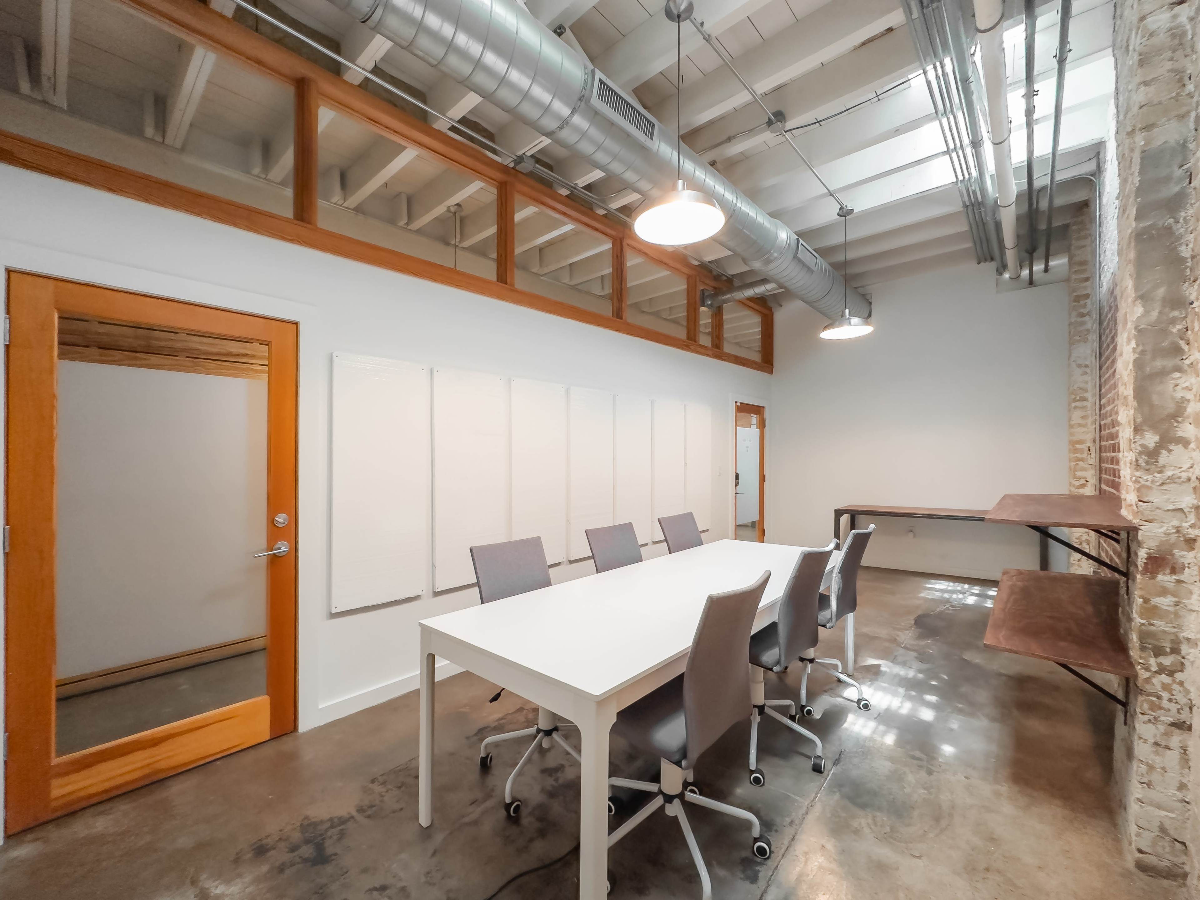 A modern conference room features a long white table surrounded by six gray chairs, with a glass-door meeting area and exposed ductwork overhead.