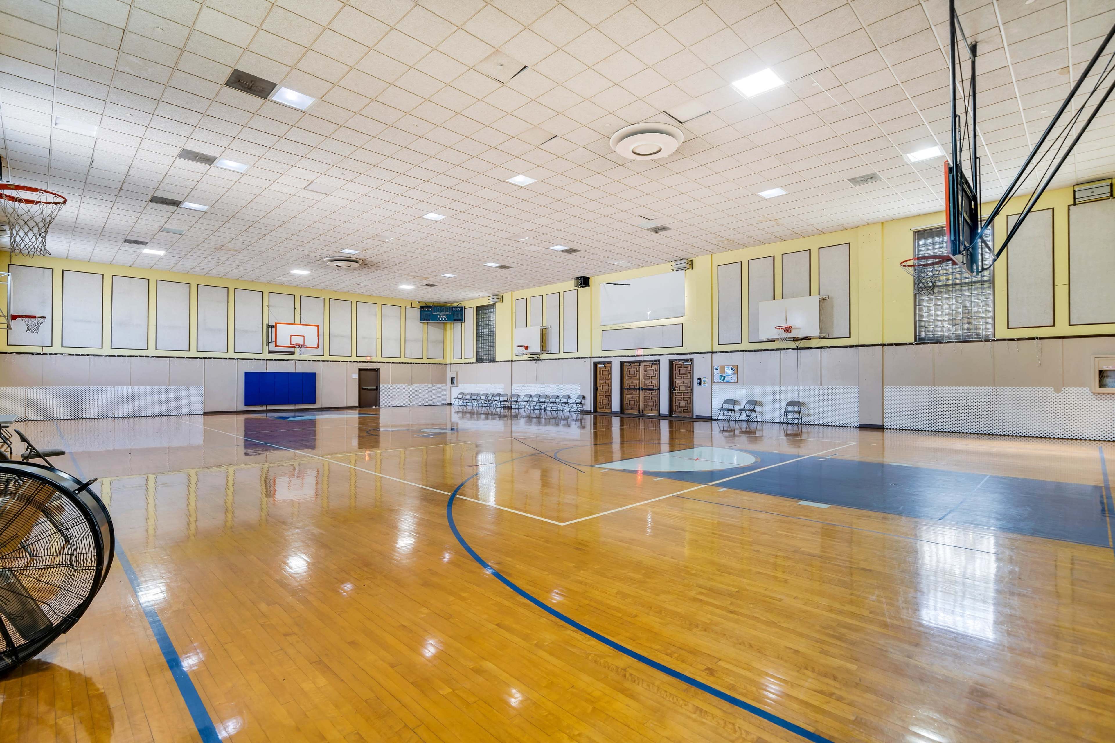 An empty gymnasium with wooden floors, basketball hoops, and folding chairs arranged along one side.