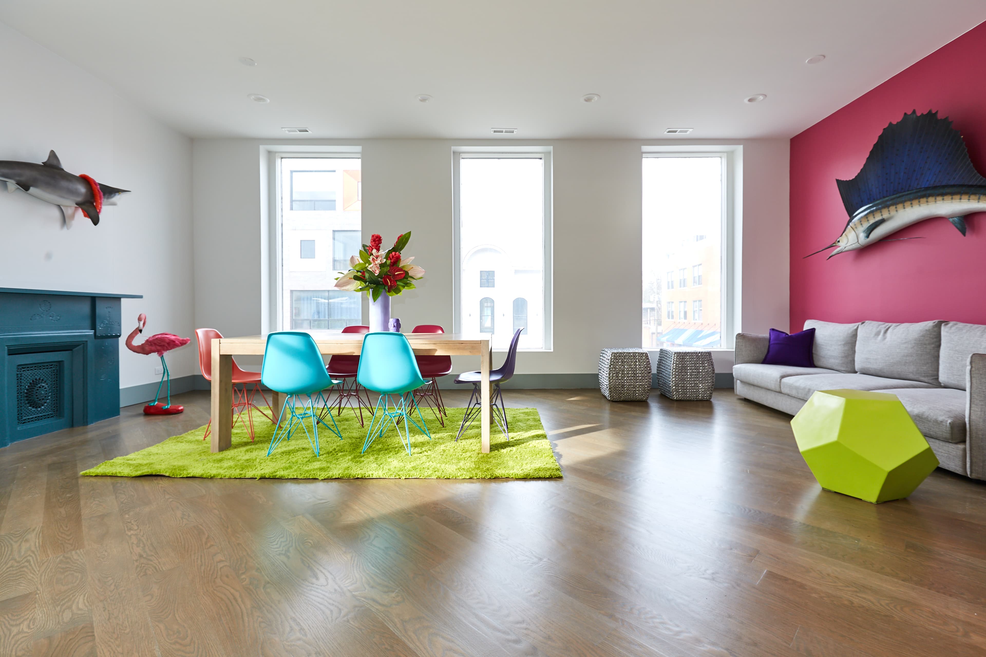 A brightly lit living space features a dining table surrounded by colorful chairs on a green rug, with large windows and decorative elements including a flamingo statue and fish mounts on the walls.