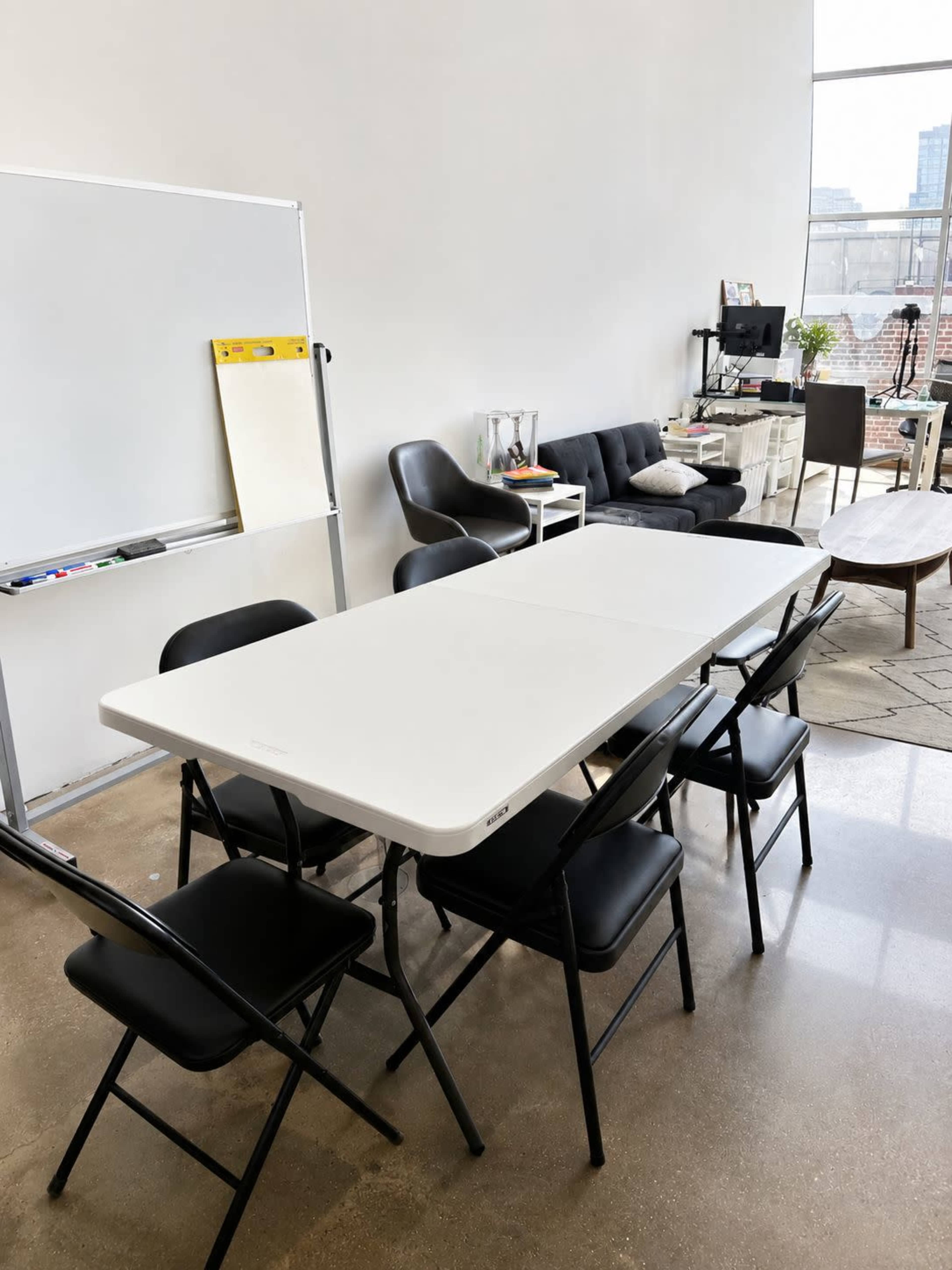 A white folding table is surrounded by six black chairs in a bright room with a whiteboard and a couch.