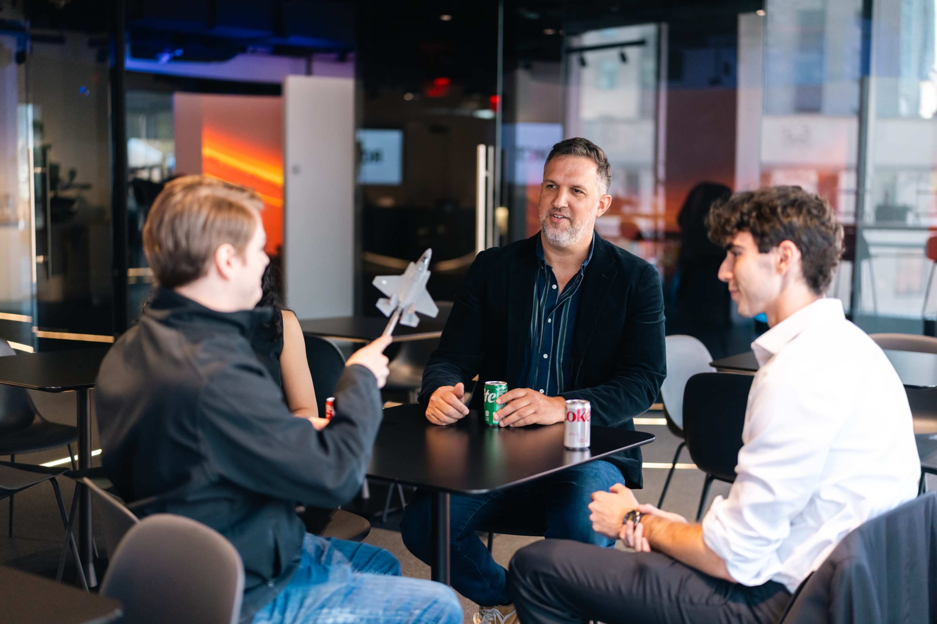 Three people are seated at a table in a modern office setting, engaging in conversation, with one person holding a toy star wand.