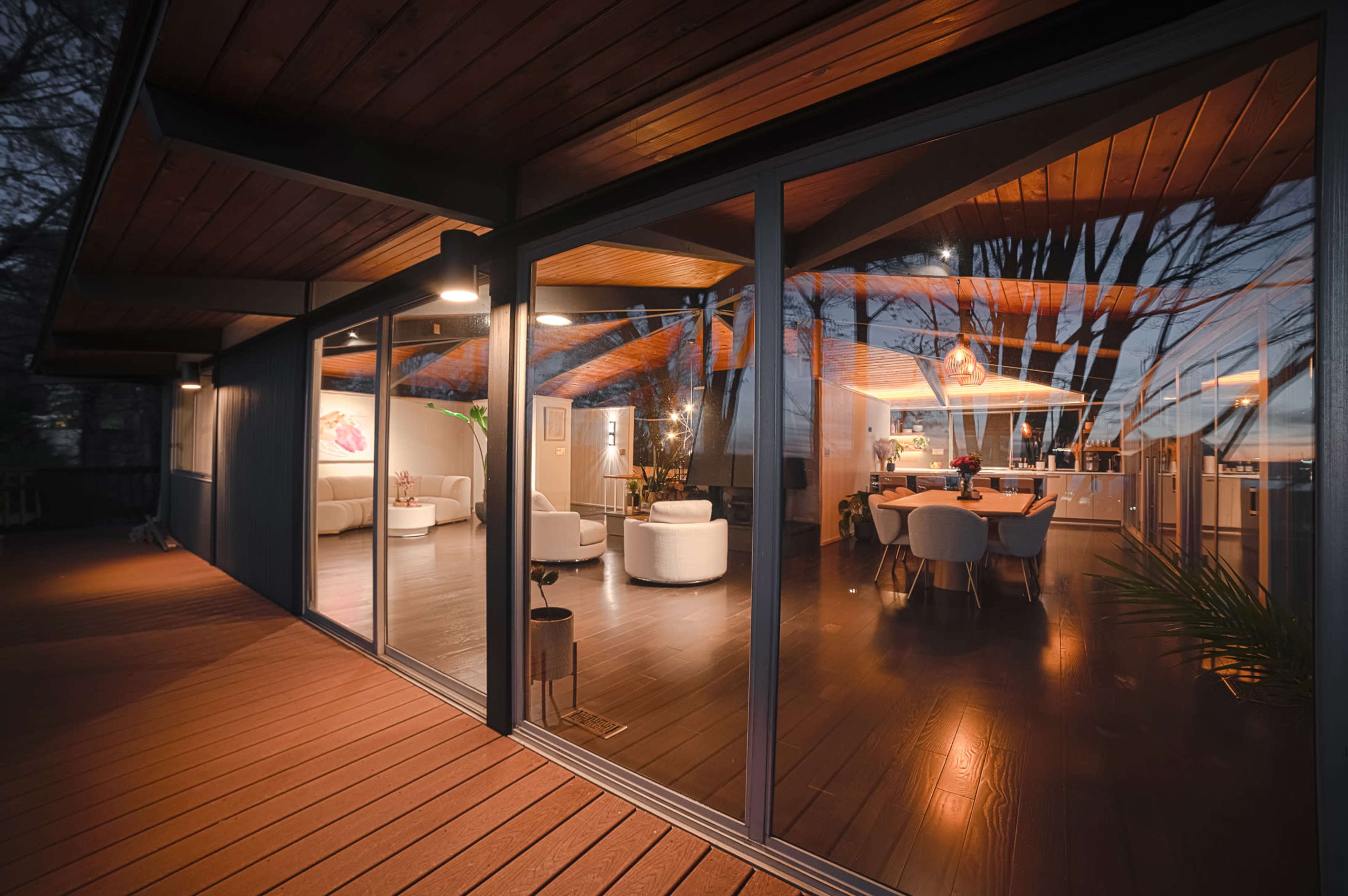 A modern house with large glass windows revealing a well-lit interior featuring a dining area and seating arrangements, surrounded by trees at dusk.