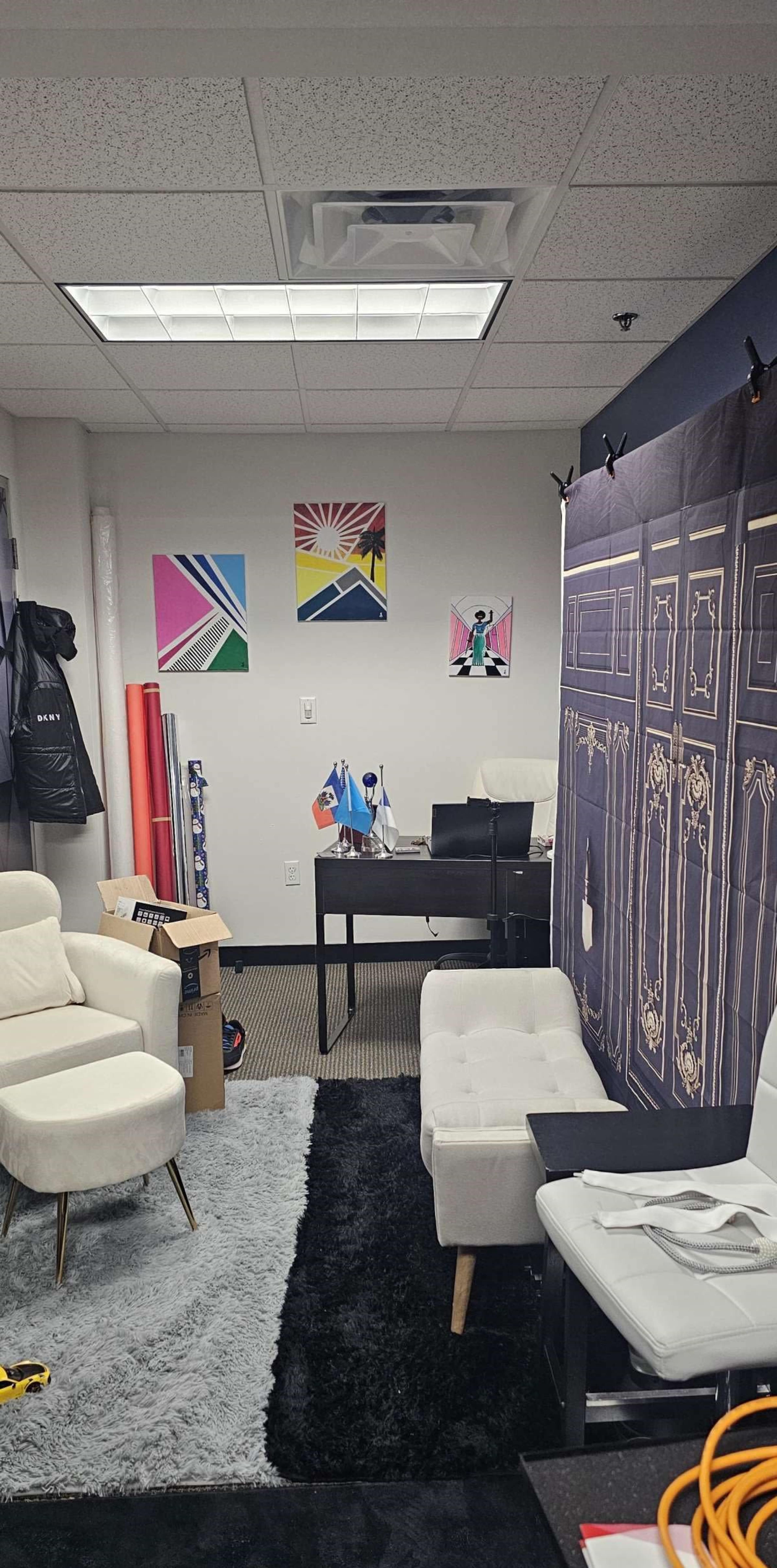 A modern office space featuring two white chairs, a desk with a chair, colorful wall art, and various supplies scattered around.