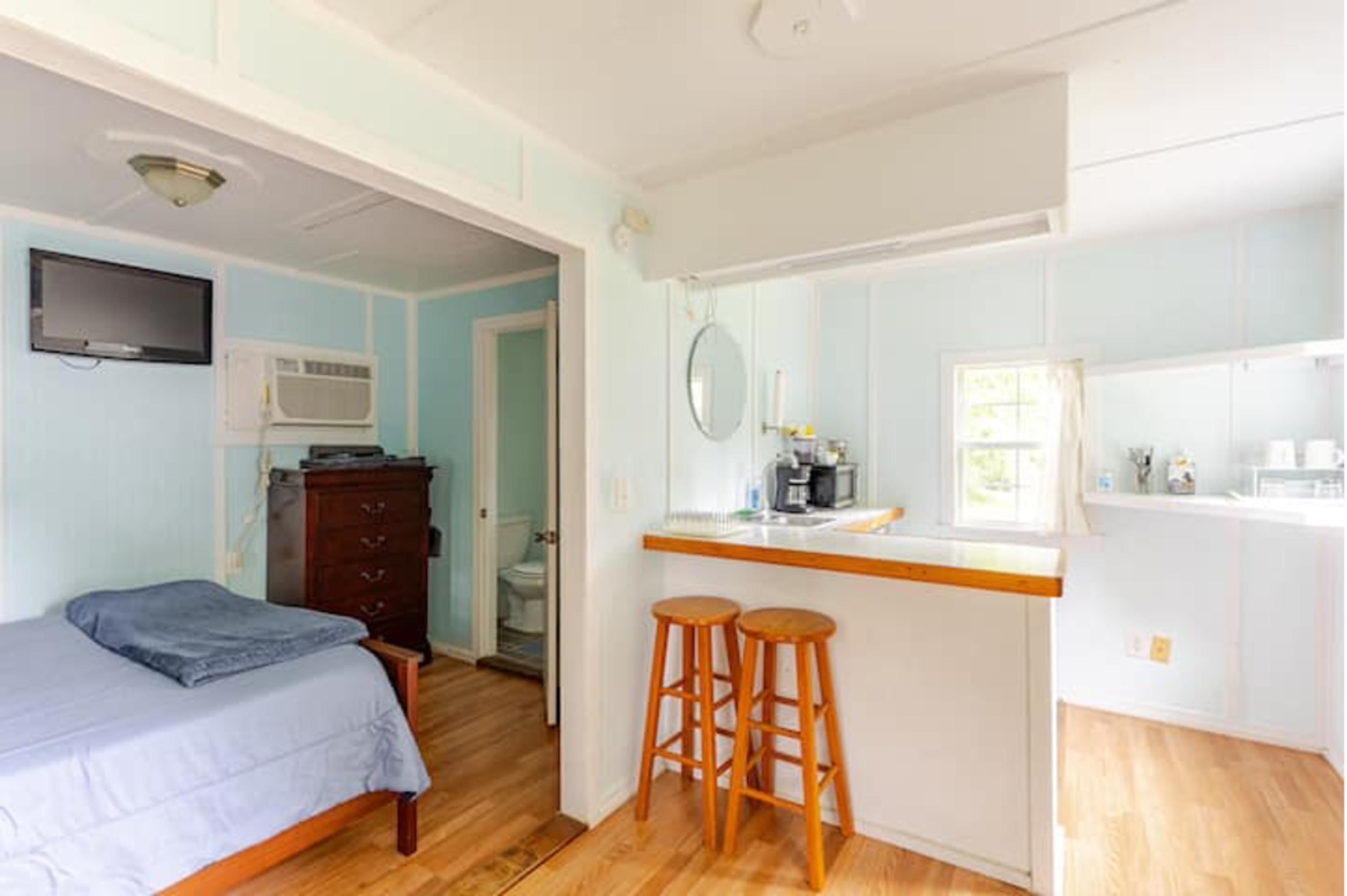 A small room with a bed, a television mounted on the wall, and a kitchenette area featuring two stools and a window.