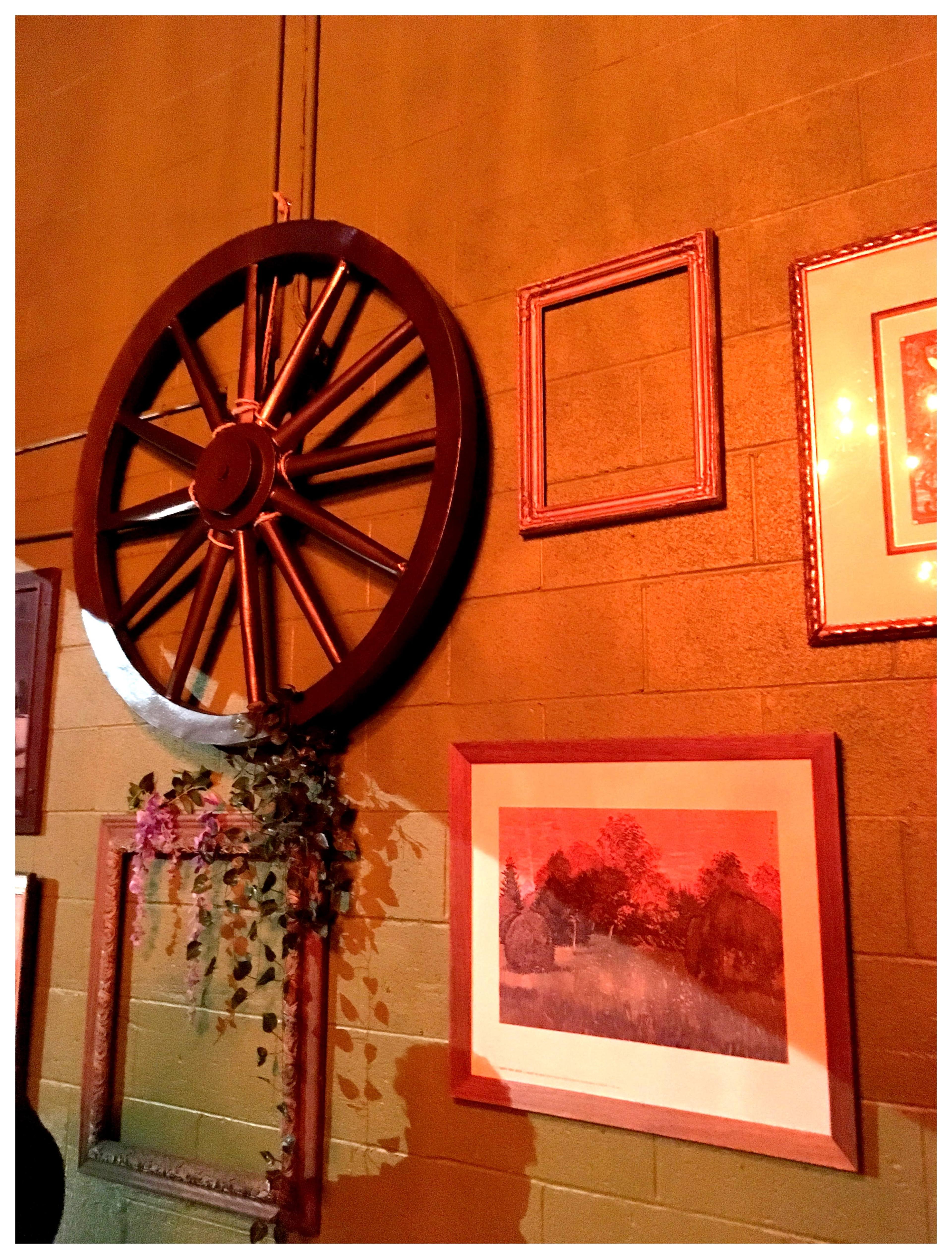 A rustic wooden wheel is mounted on a textured wall, surrounded by various empty frames and a framed painting depicting a landscape.