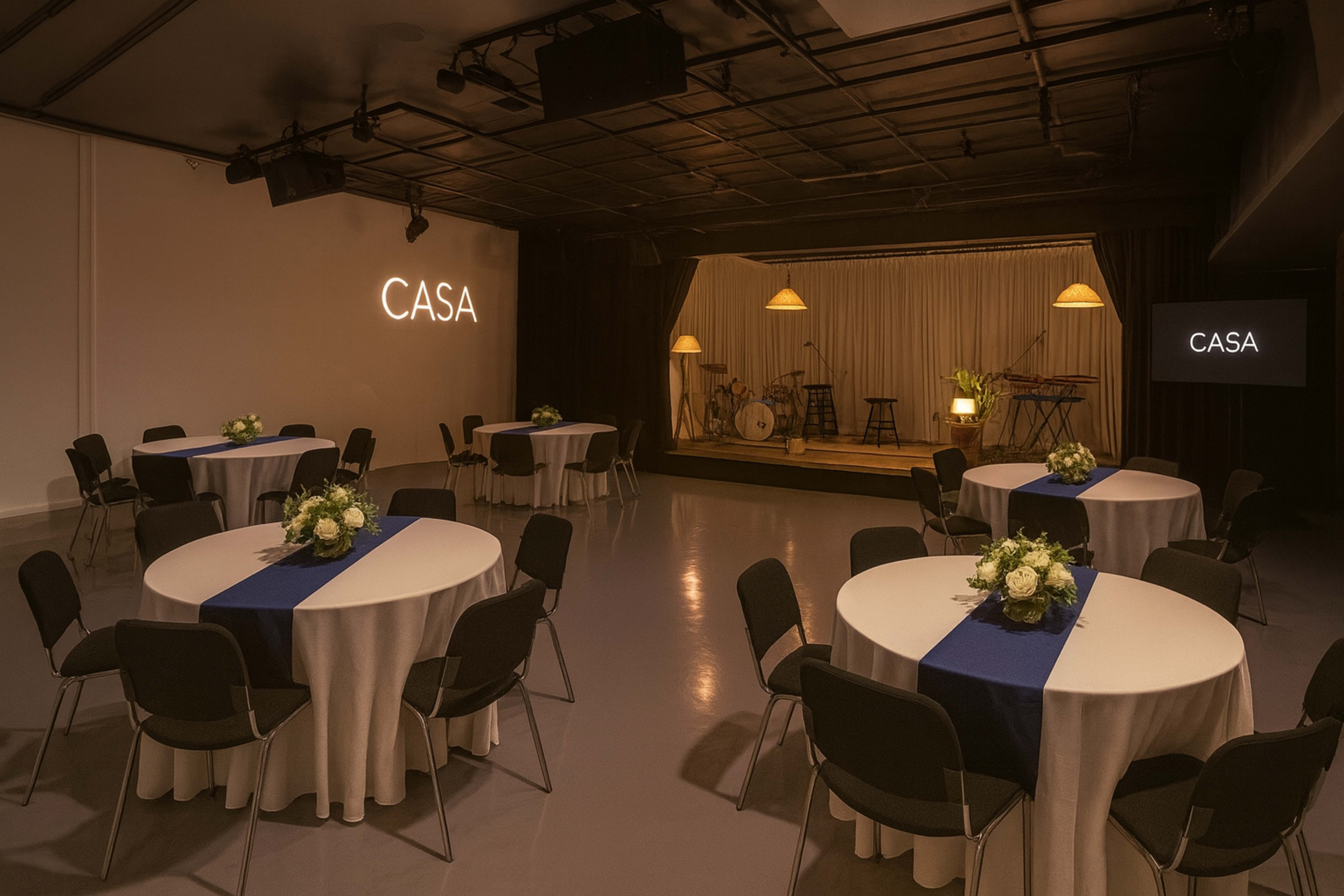 A modern event space features round tables with white tablecloths and blue runners, arranged around a small stage with musical instruments.