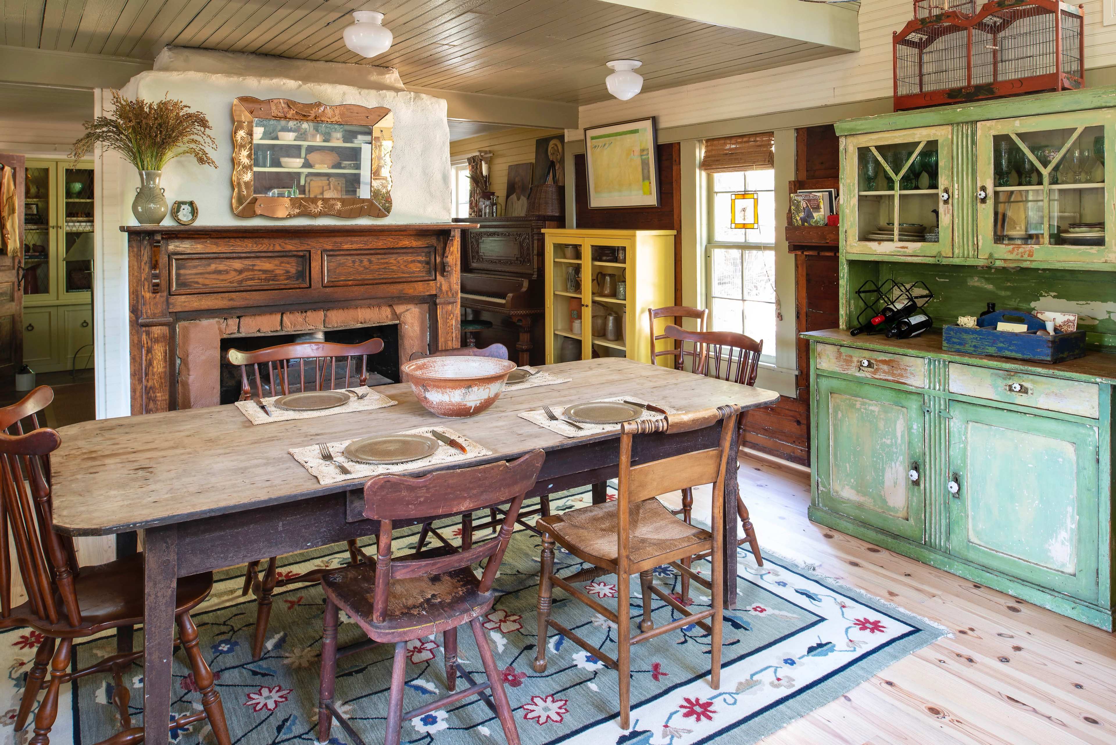 A rustic dining room features a long wooden table surrounded by mismatched chairs, with a fireplace and a green cabinet in the background.
