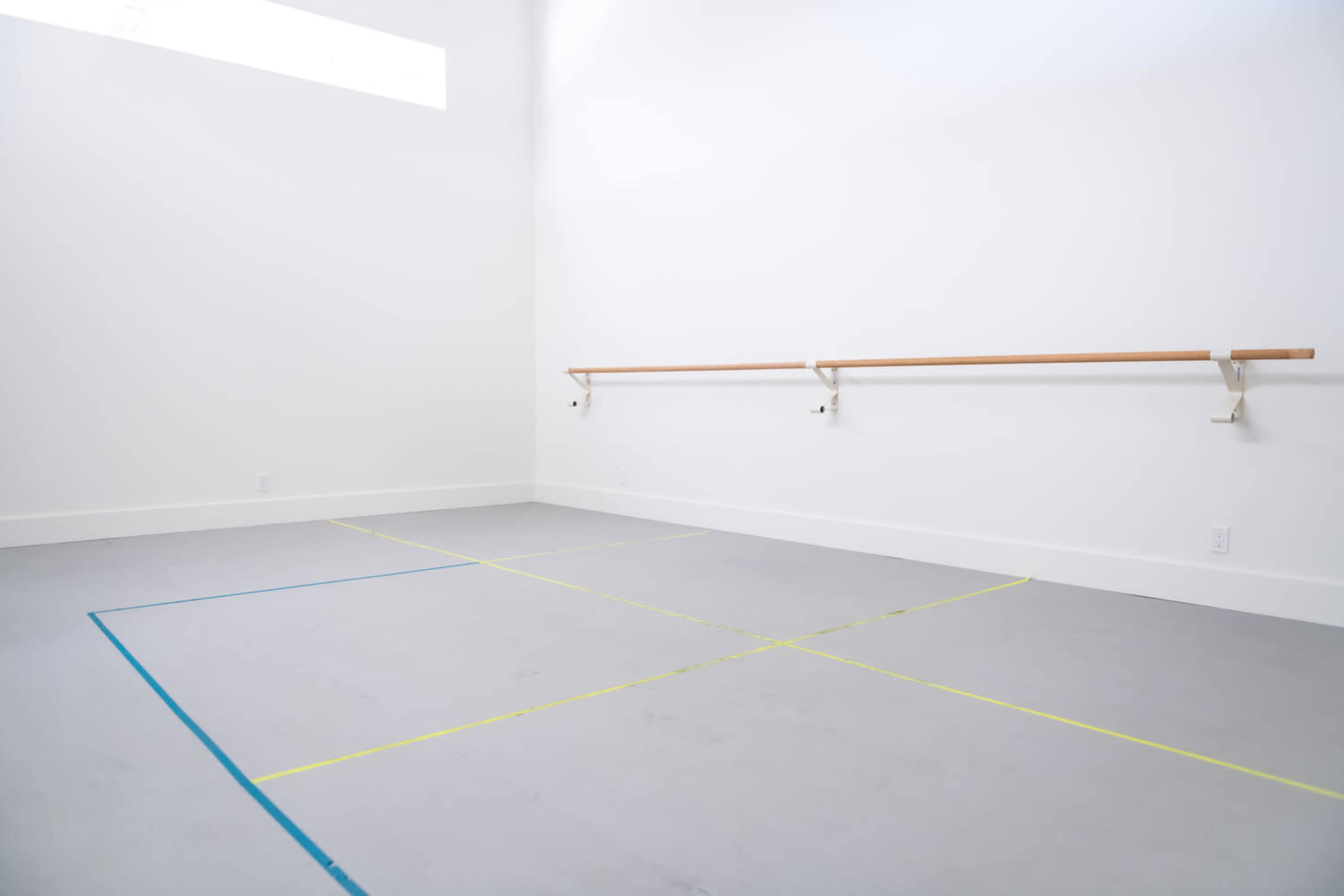 A dance studio with wooden ballet bars mounted on a white wall and a marked floor featuring blue and yellow lines.