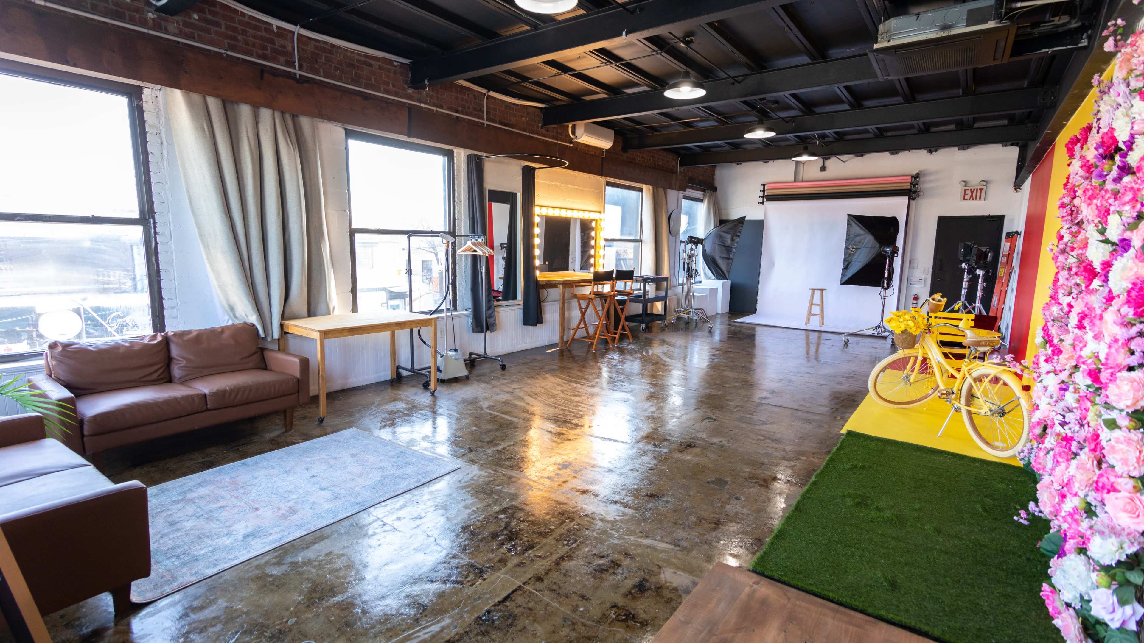 A spacious studio interior with large windows, a seating area, a wooden table, and a backdrop featuring a colorful floral arrangement.