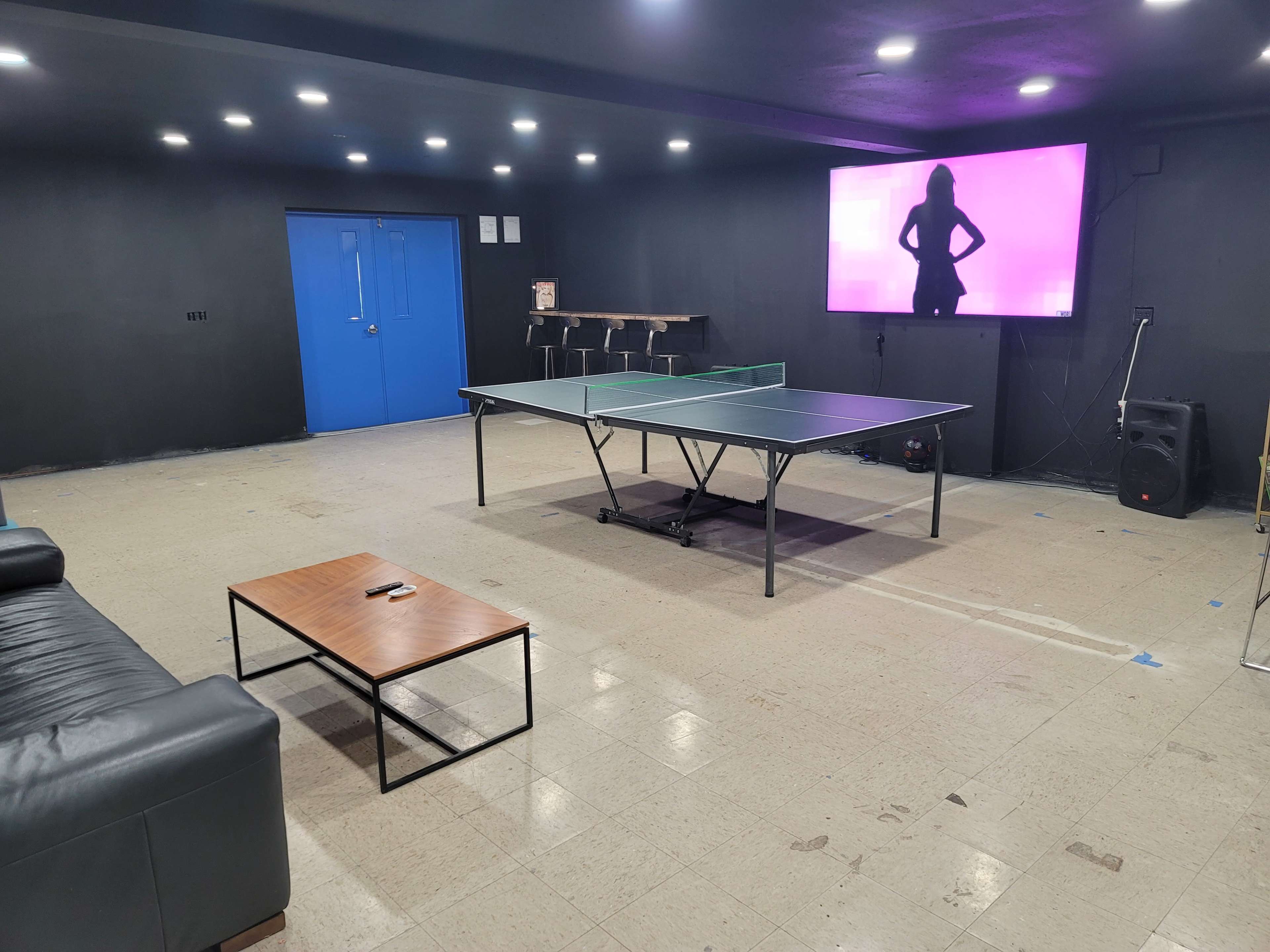 A spacious room features a ping pong table, a coffee table, a black sofa, and a large screen displaying a silhouette against a pink background.