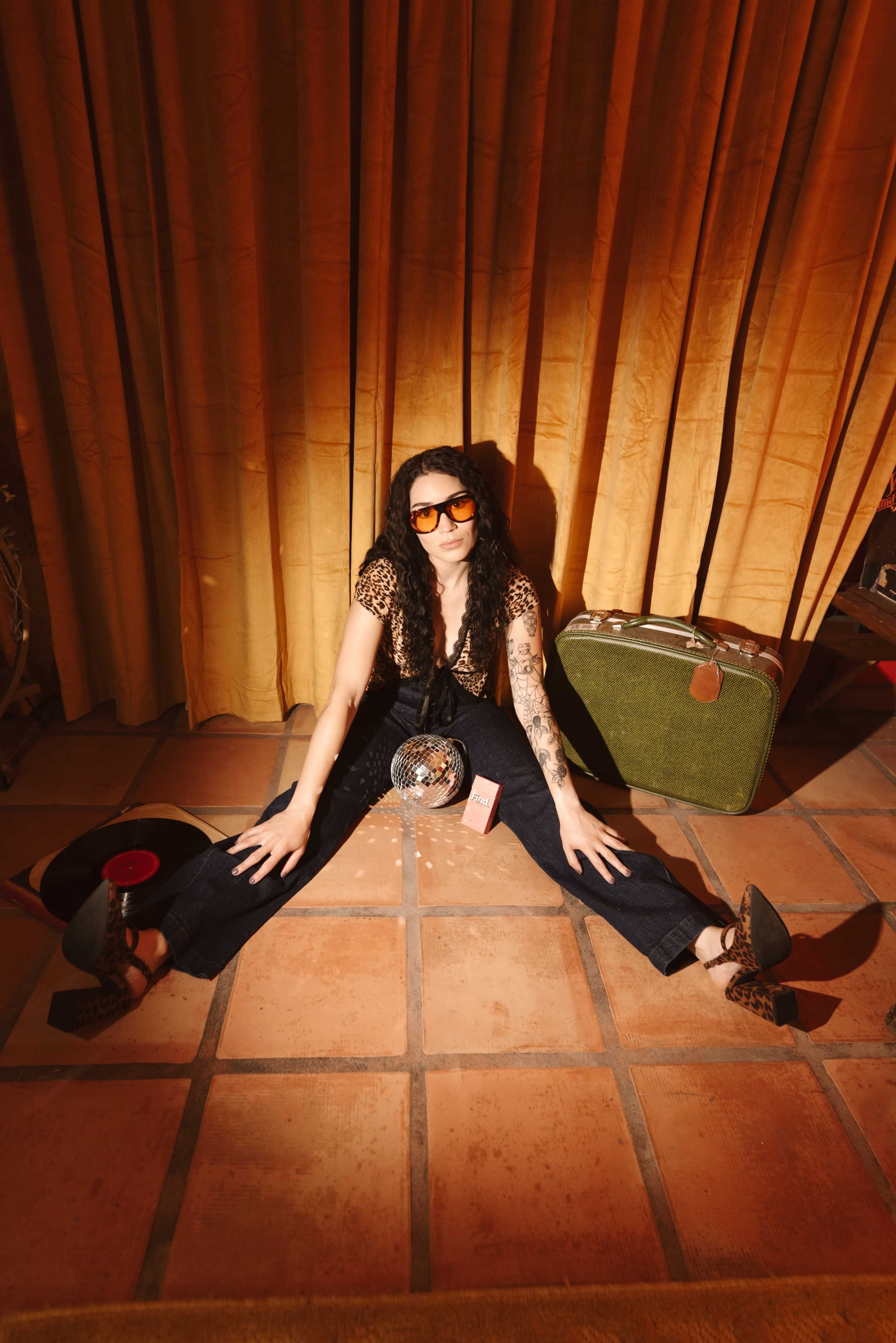 A woman in sunglasses sits on a tiled floor, with her legs spread and various vintage items, including a suitcase and a disco ball, arranged around her.