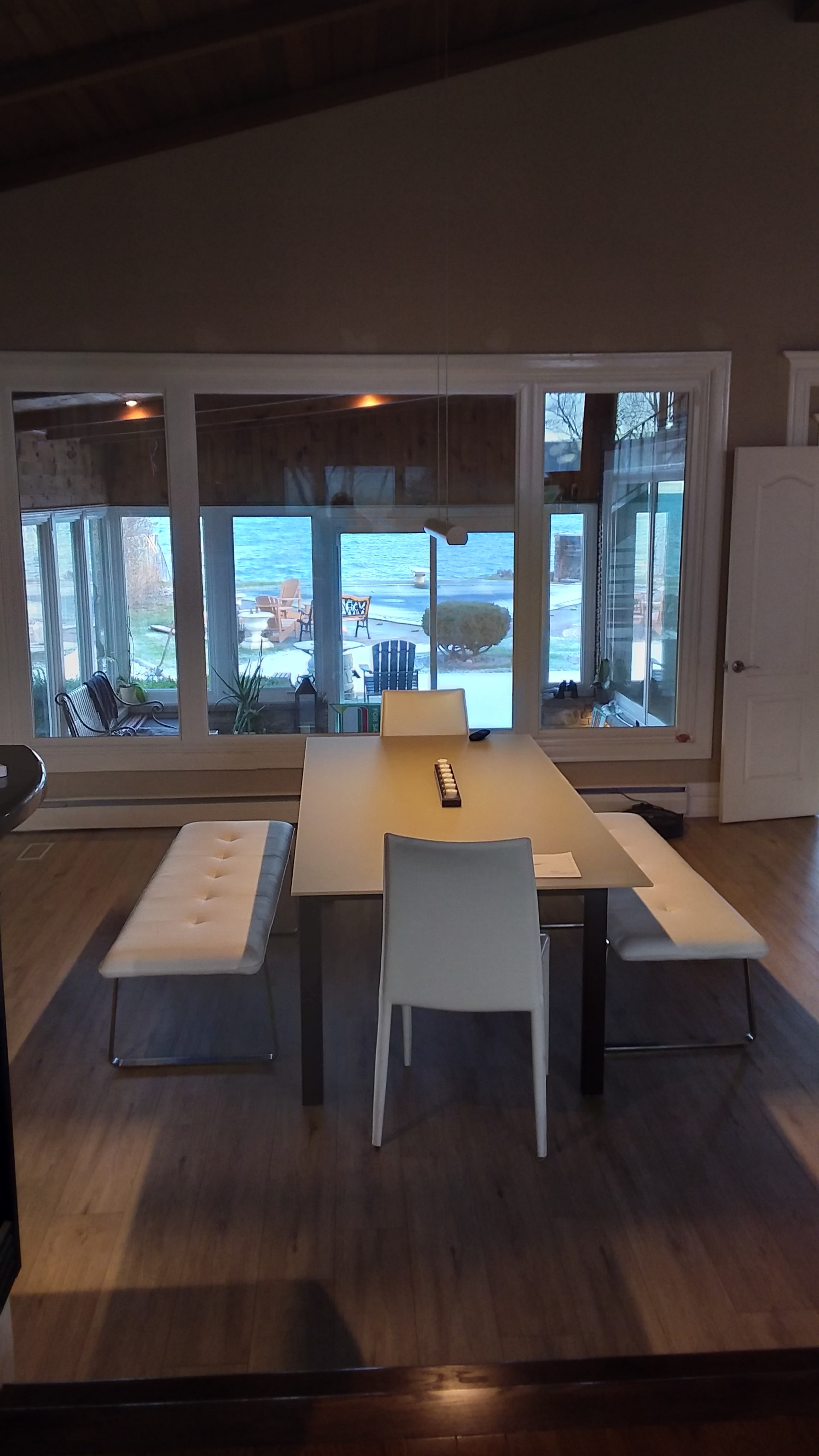 A dining area with a rectangular table and white chairs, overlooking large windows with a view of water and outdoor seating.