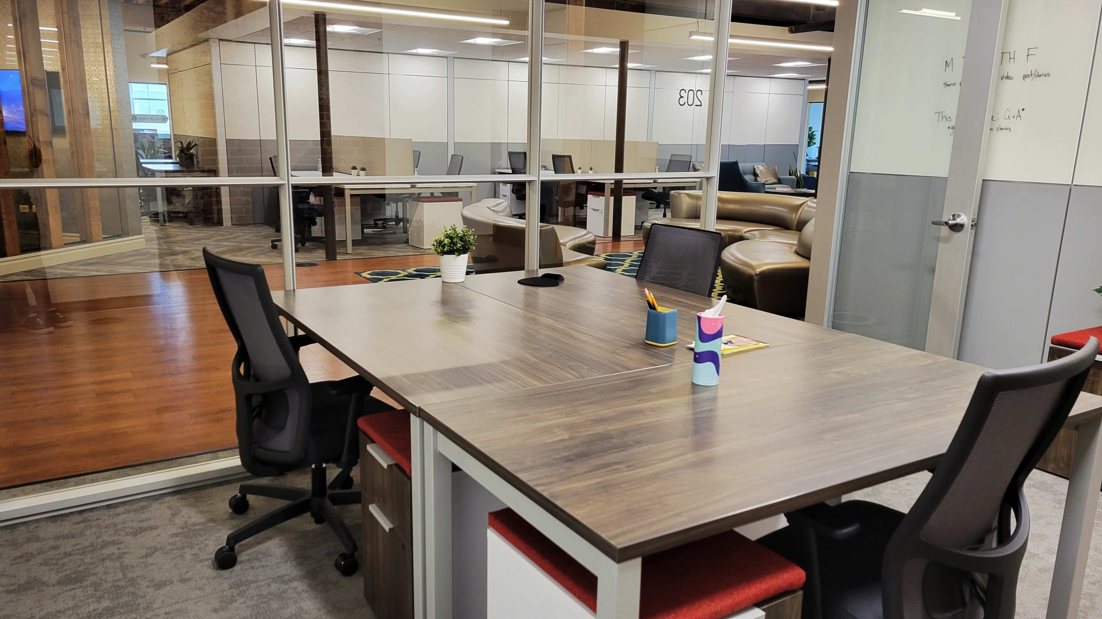 The image shows a modern office space with a large wooden table at the forefront, flanked by two black chairs and surrounded by glass-walled workstations in the background.