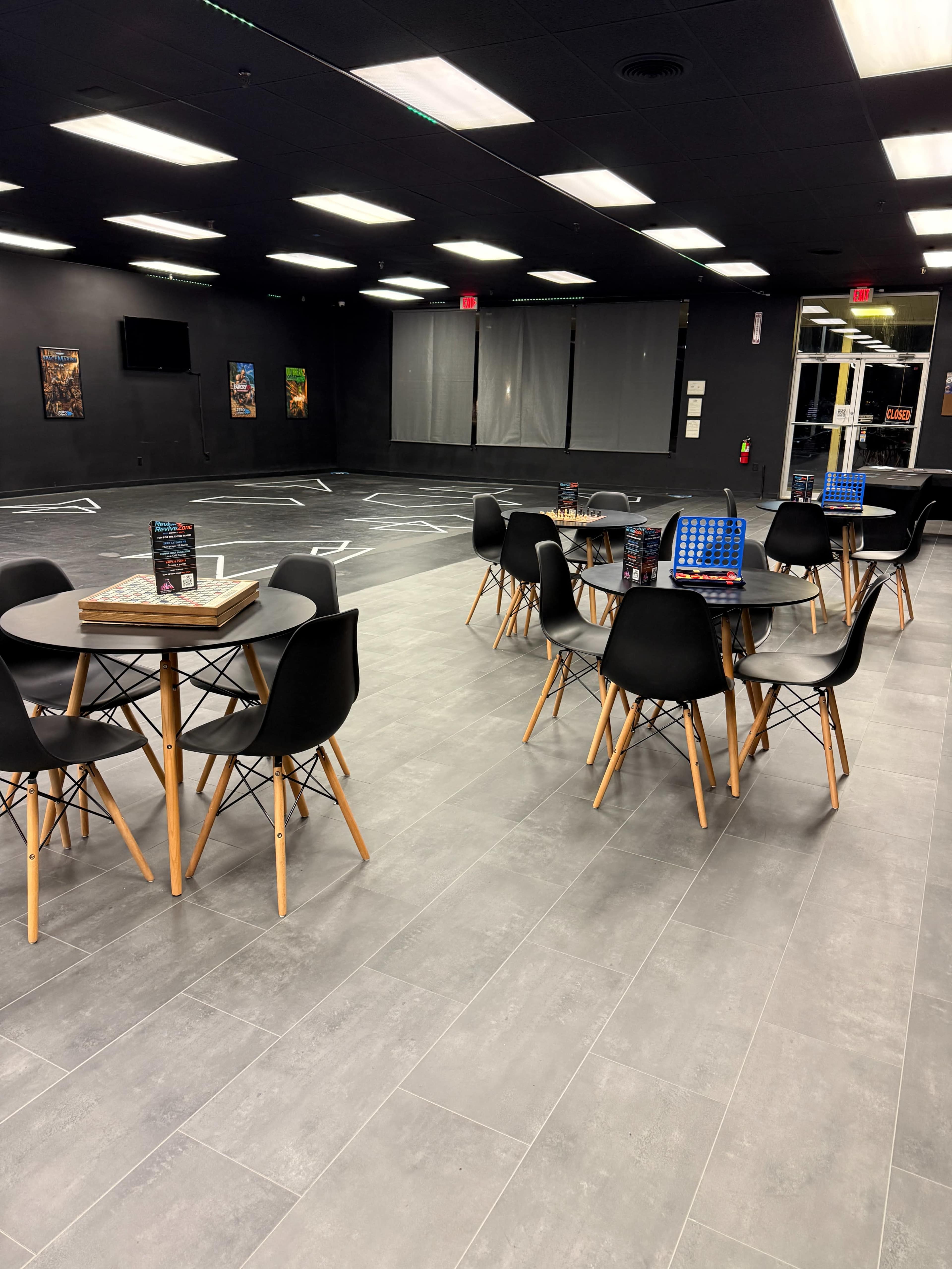 The image shows a spacious indoor area with several round tables and black chairs, illuminated by fluorescent lights, and featuring a dark wall backdrop.