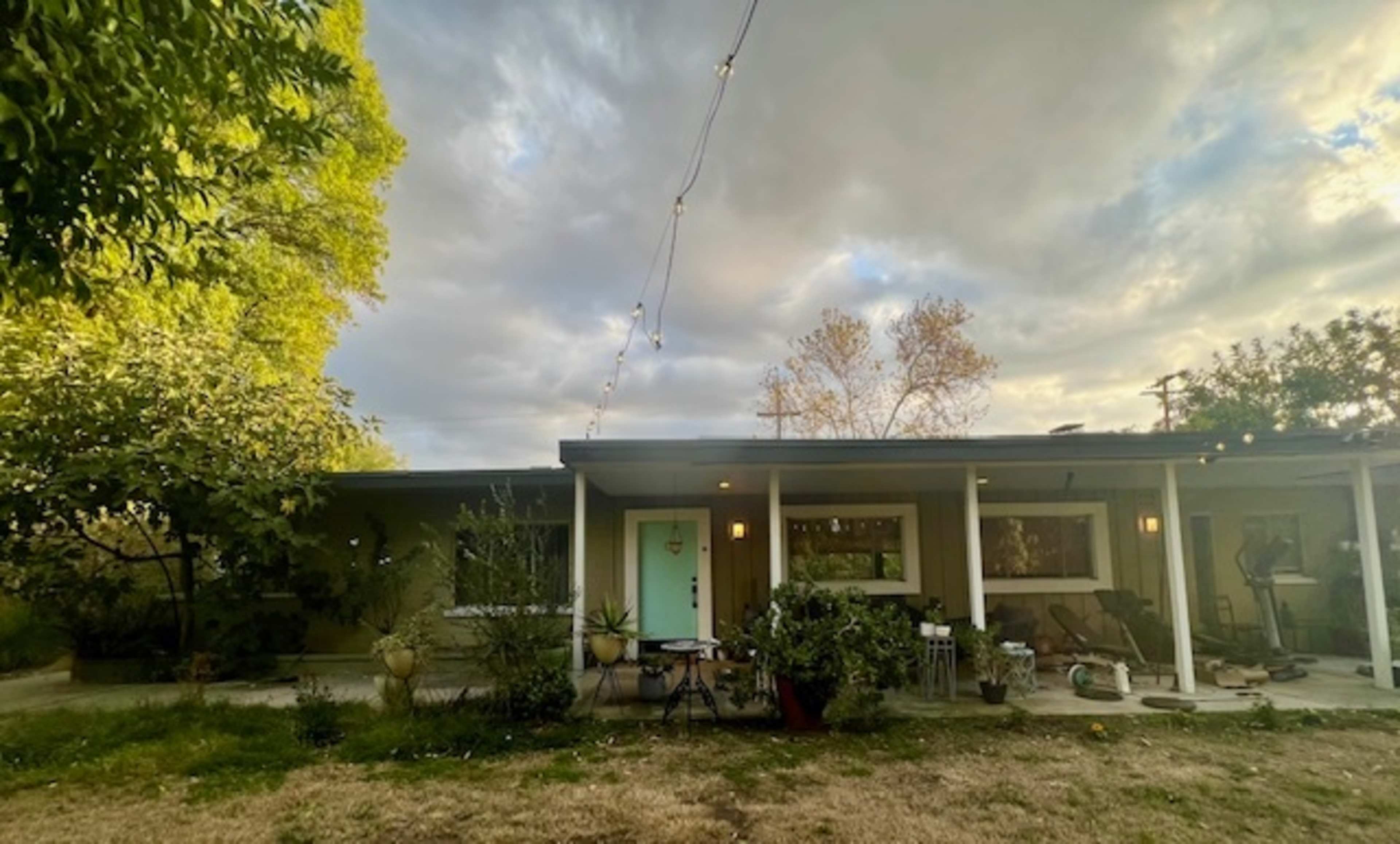 A single-story house with a teal front door is surrounded by potted plants and has string lights overhead under a cloudy sky.