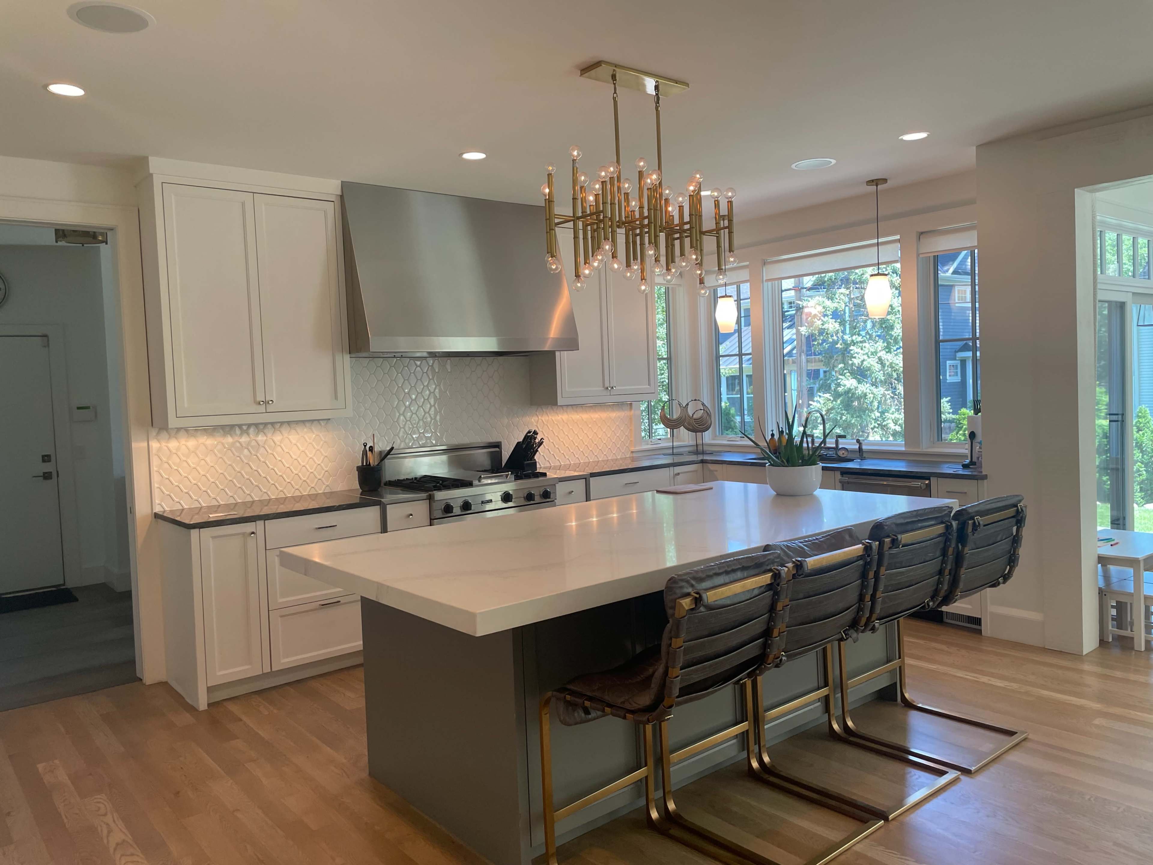 The image shows a modern kitchen featuring white cabinetry, a large island with bar seating, a stainless steel range hood, and bright windows overlooking a garden.