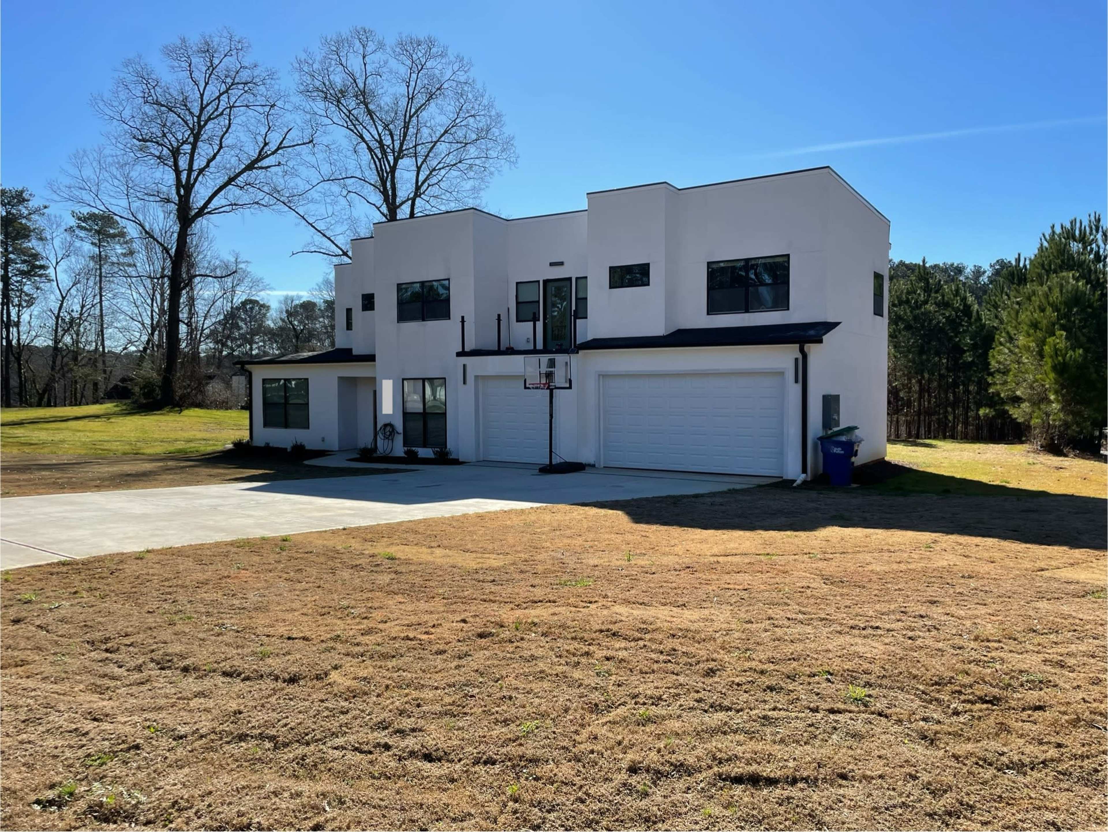 A modern two-story house with a flat roof and large windows is situated on a grassy lot, featuring a basketball hoop and a driveway.