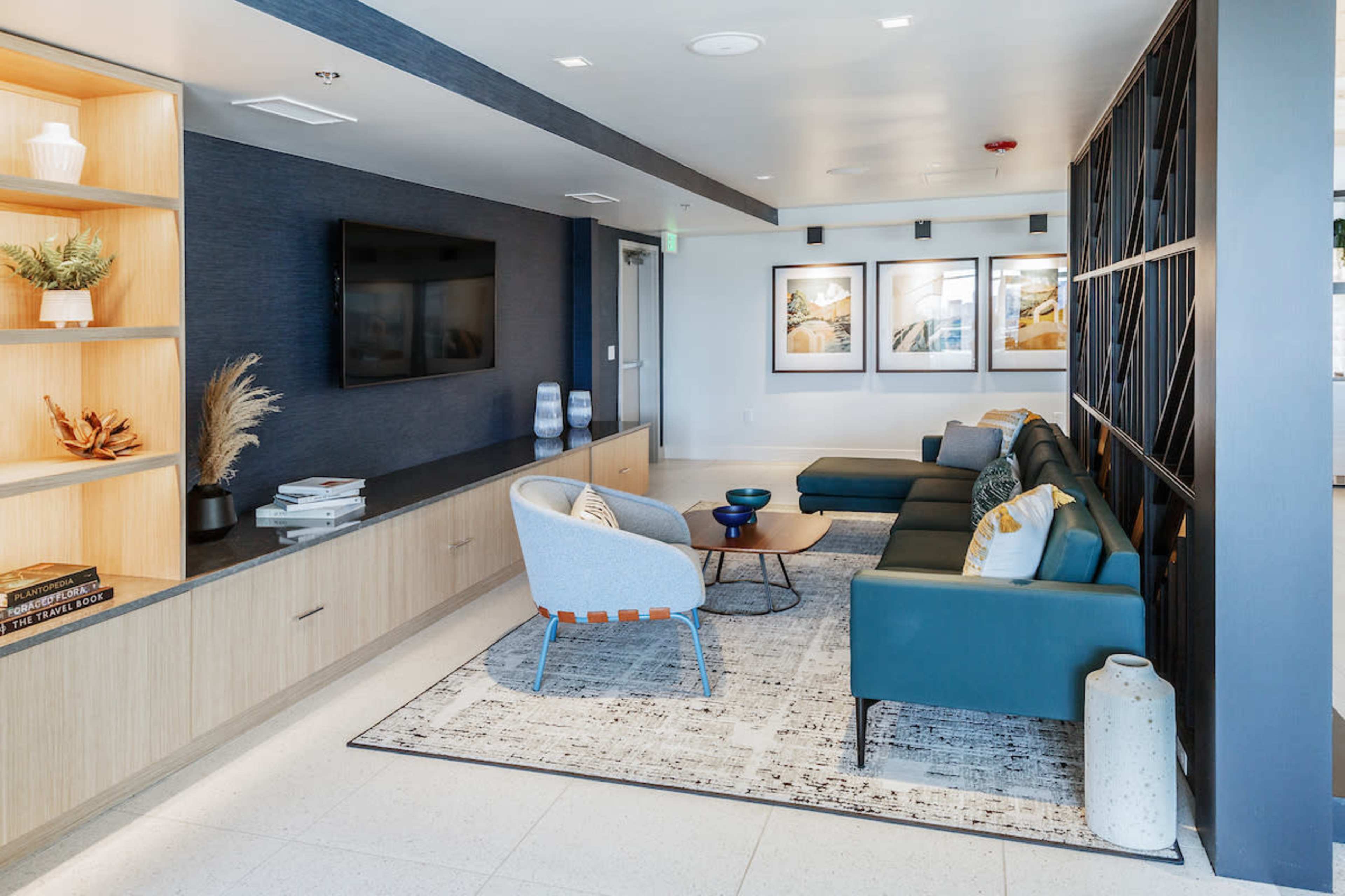 A modern lounge area featuring a blue sectional sofa, a minimalist wooden cabinet, a flat-screen TV mounted on the wall, and framed artwork.