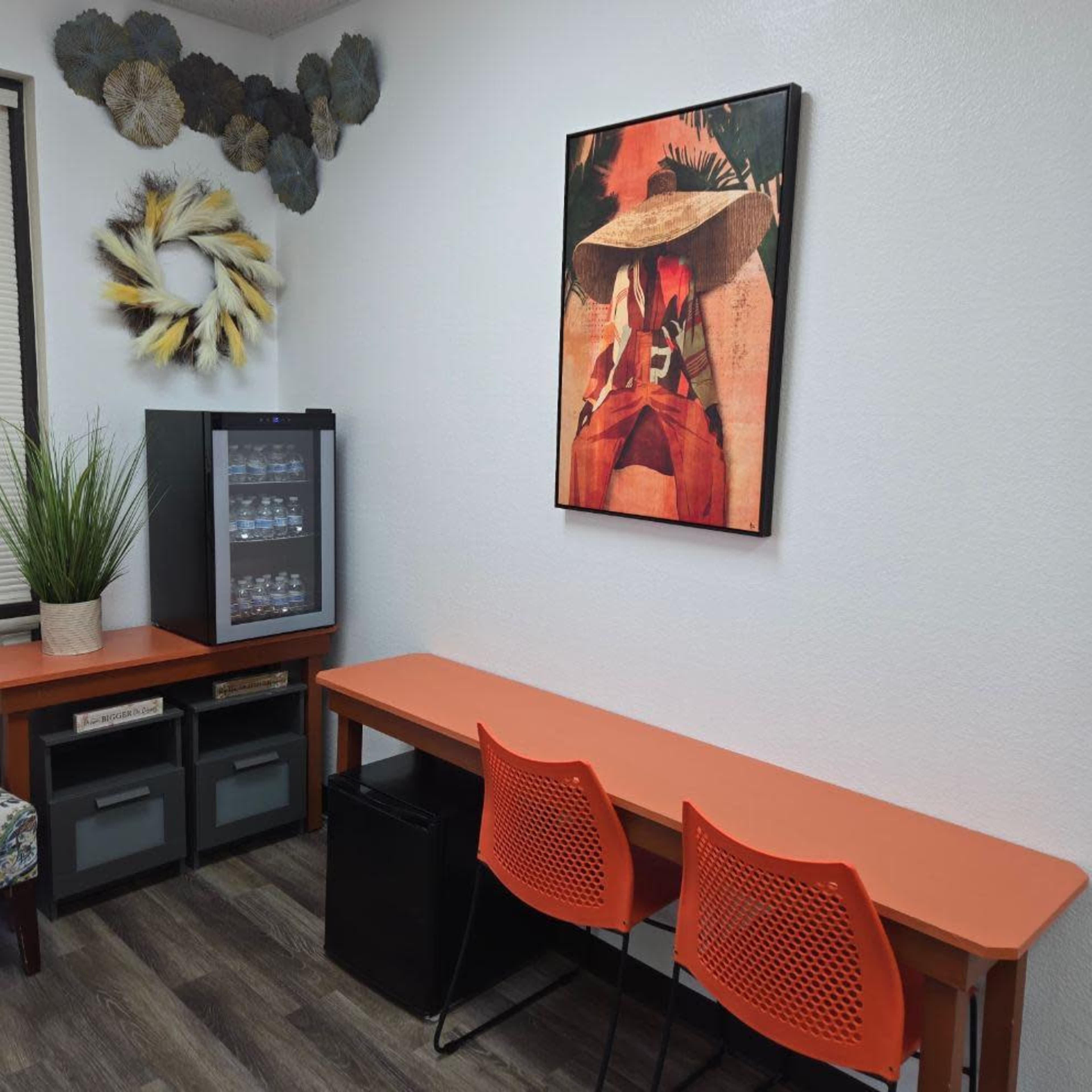 The image shows a small room with an orange table and chairs, a black mini fridge, and a framed artwork featuring a person in a hat on the wall.