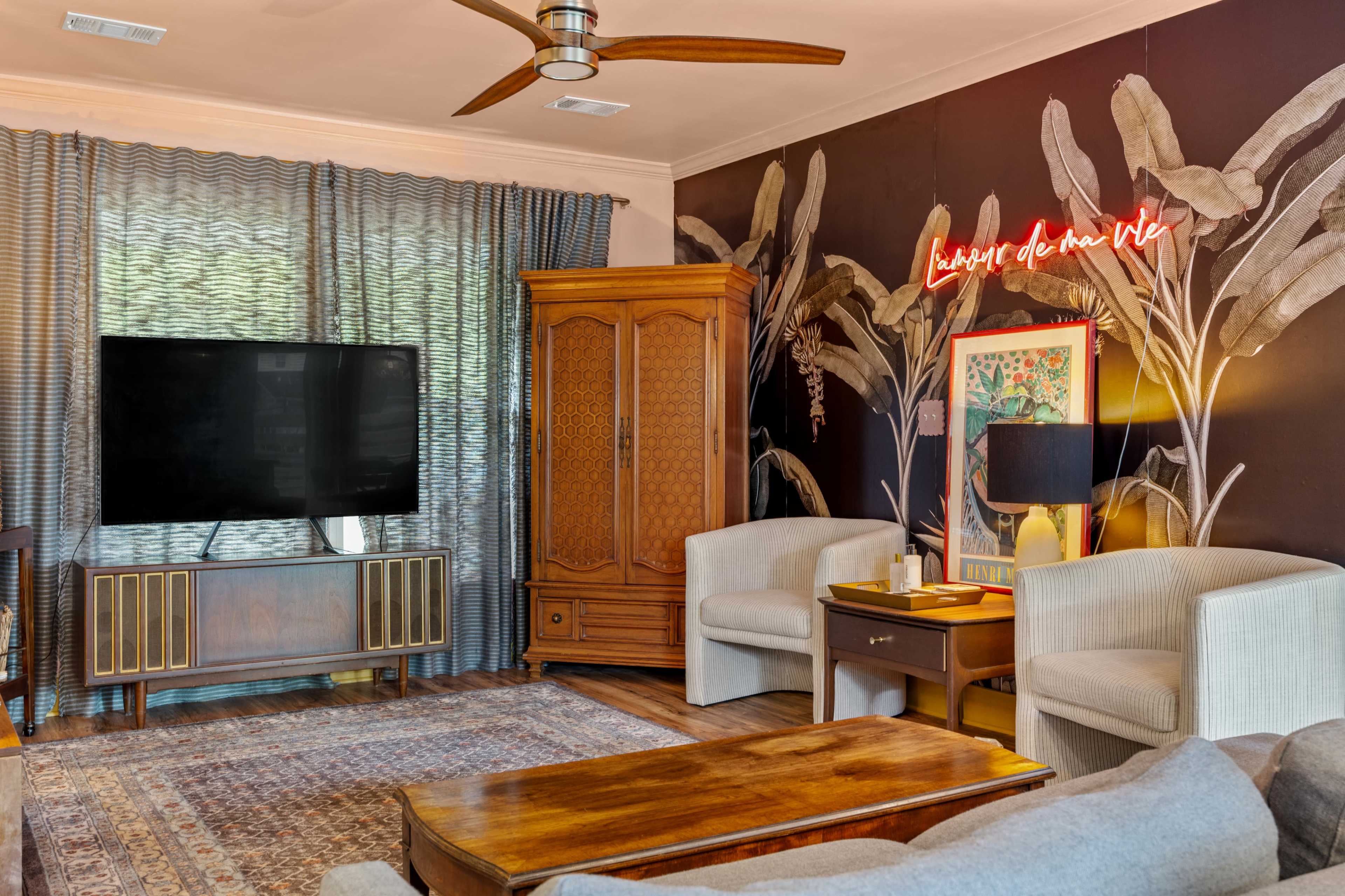 The image shows a living room with a large television, wooden furniture, and decorative wall art featuring tropical leaves.
