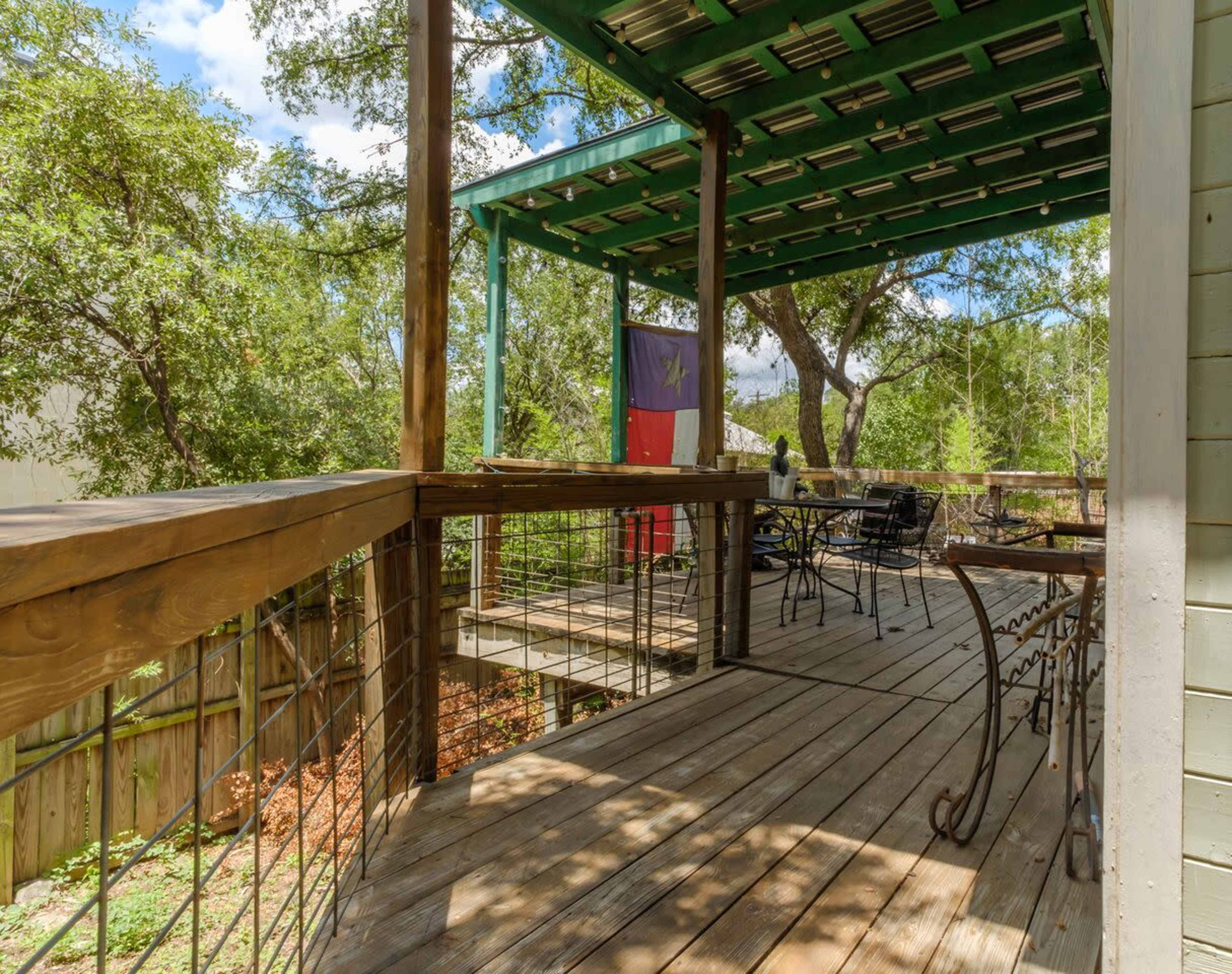 A wooden deck with a green roof, featuring a flag and outdoor furniture surrounded by trees.
