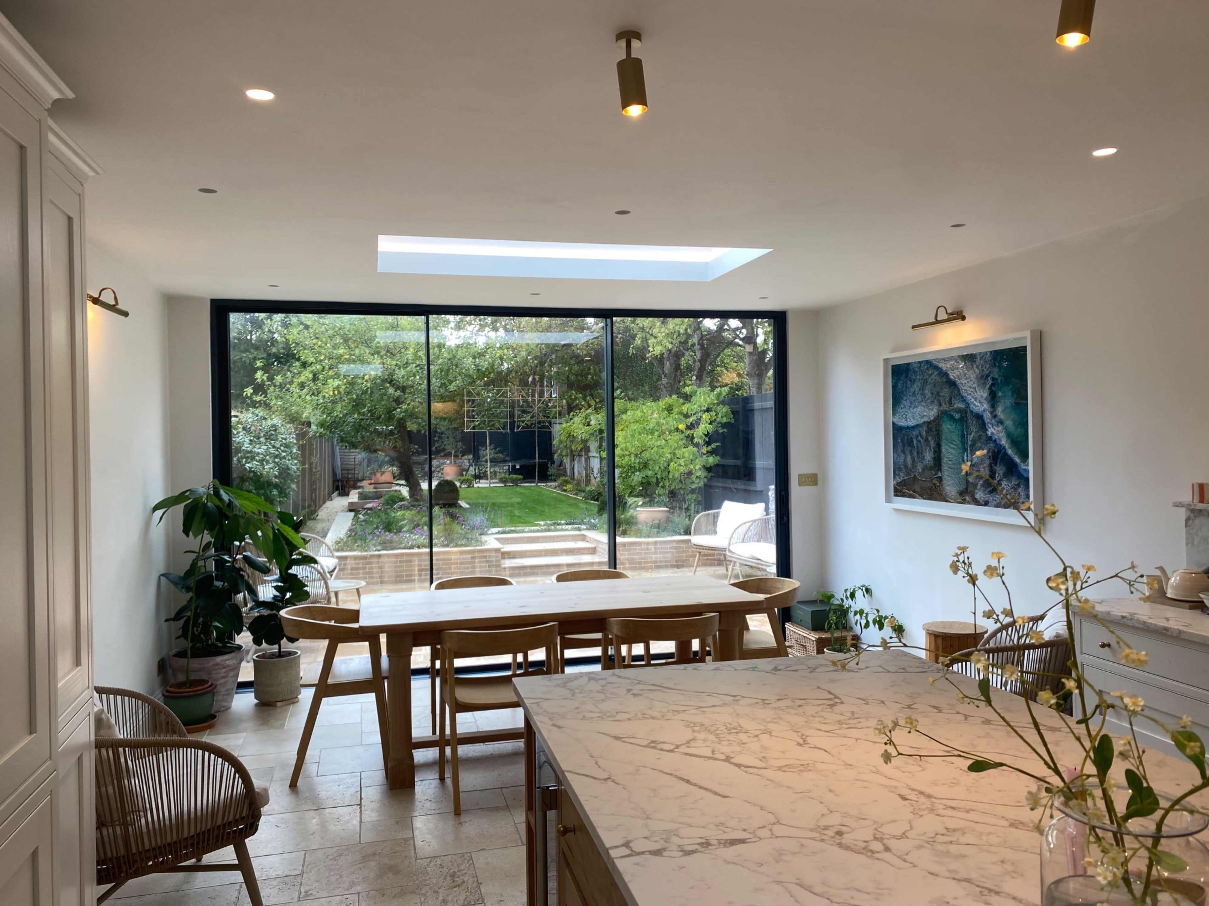 The image shows a modern kitchen and dining area with a long wooden table, surrounded by chairs, and a large glass window overlooking a garden.