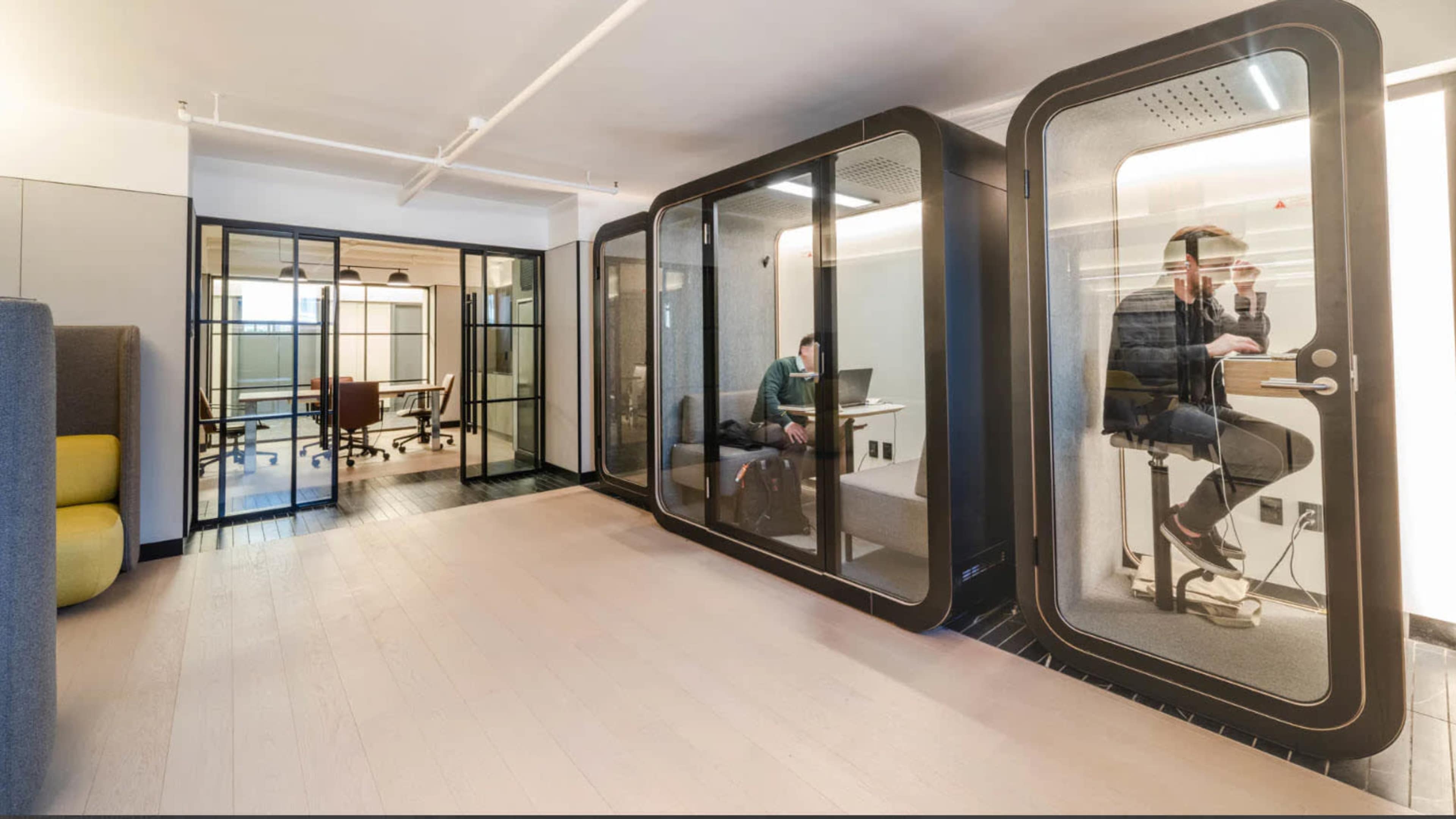 A modern office space featuring glass-enclosed meeting pods and a collaborative area with desks and chairs.