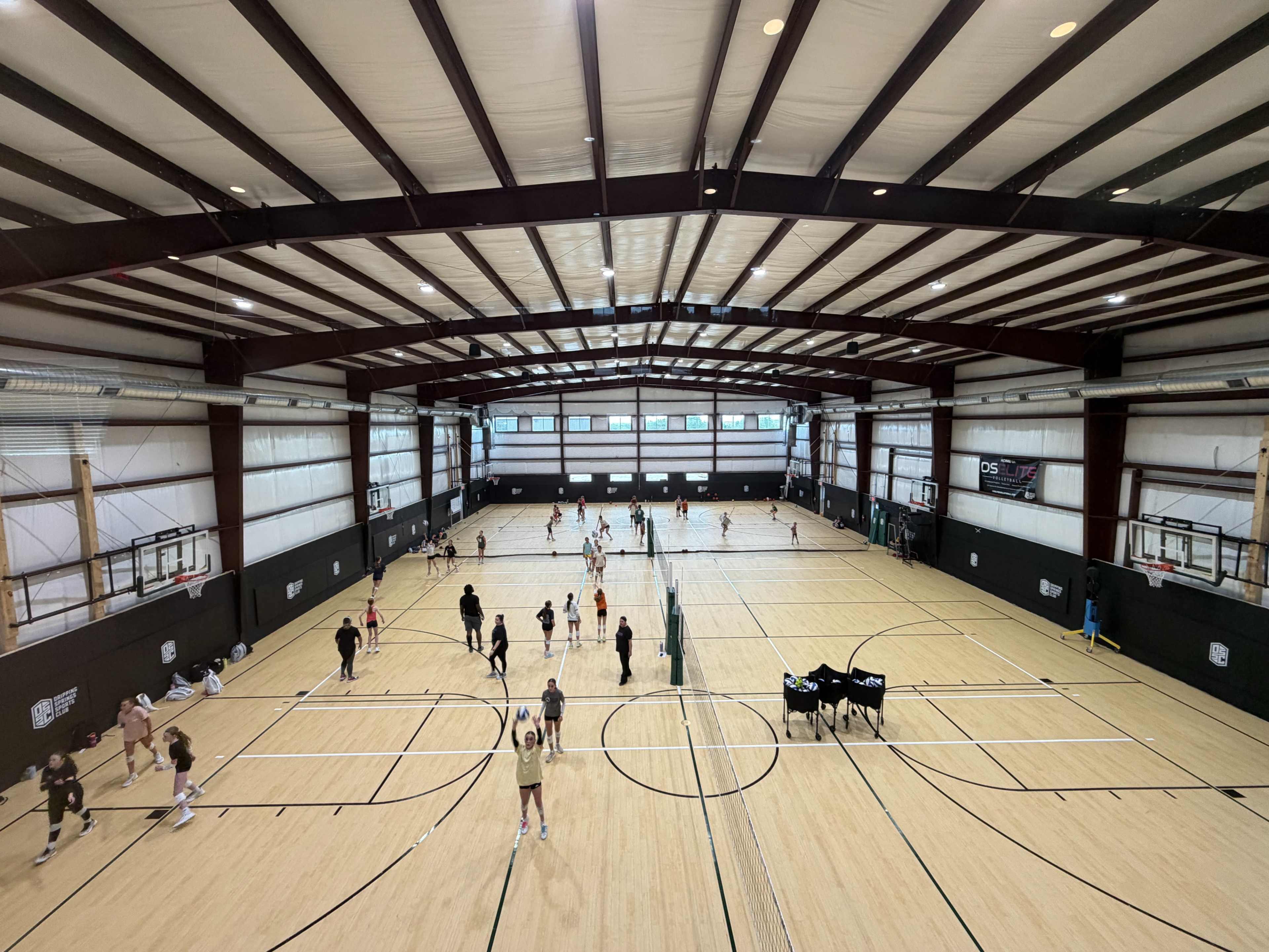 The image shows a large indoor sports facility with multiple basketball courts, where various players are engaged in practice and warm-up activities.