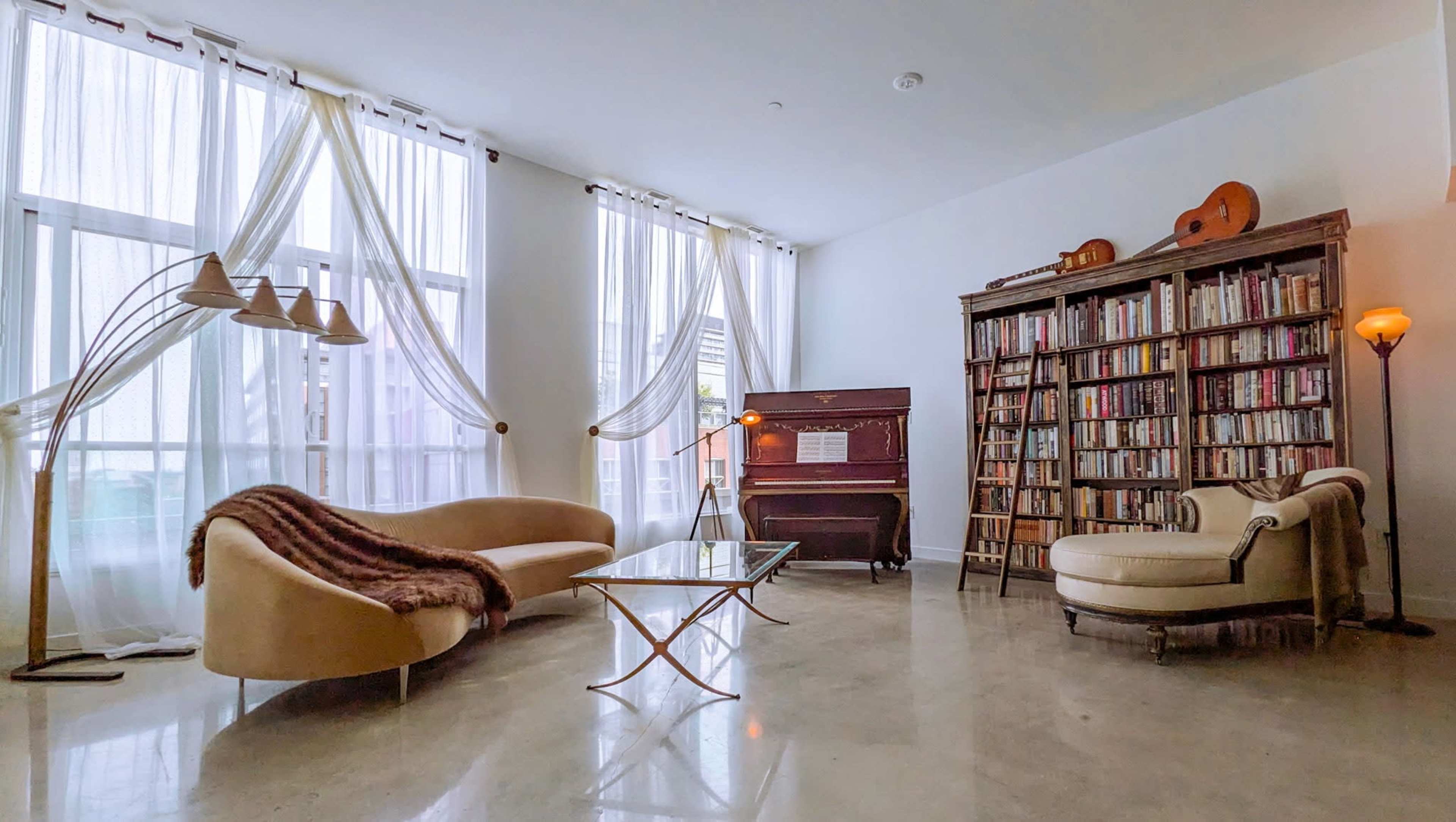 The room features a modern seating arrangement with a curved sofa, a glass coffee table, an upright piano, and a tall bookshelf filled with books, all illuminated by natural light filtering through large curtains.
