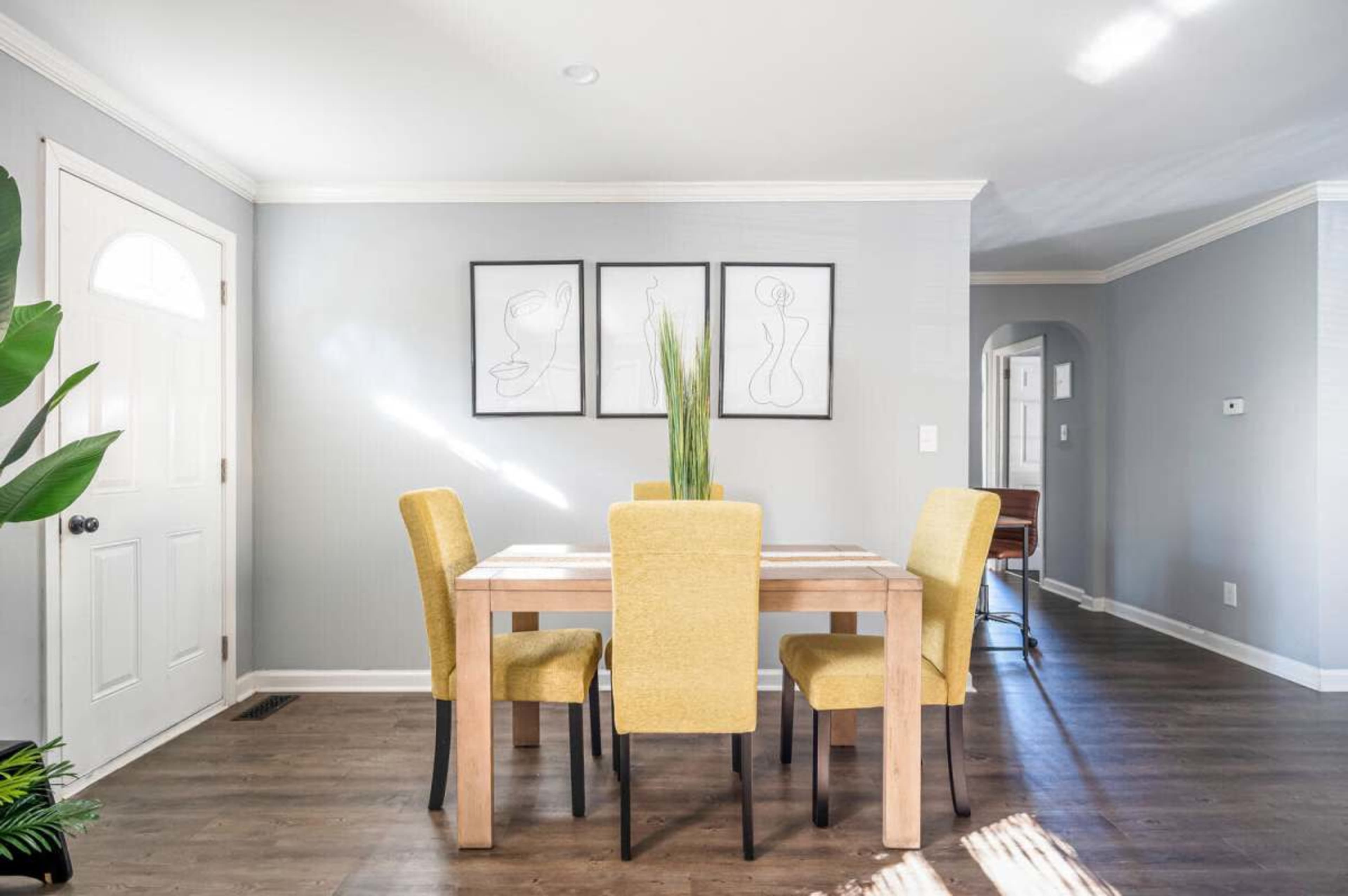 The image shows a well-lit dining area with a wooden table surrounded by four yellow chairs, accompanied by framed abstract art on the walls.