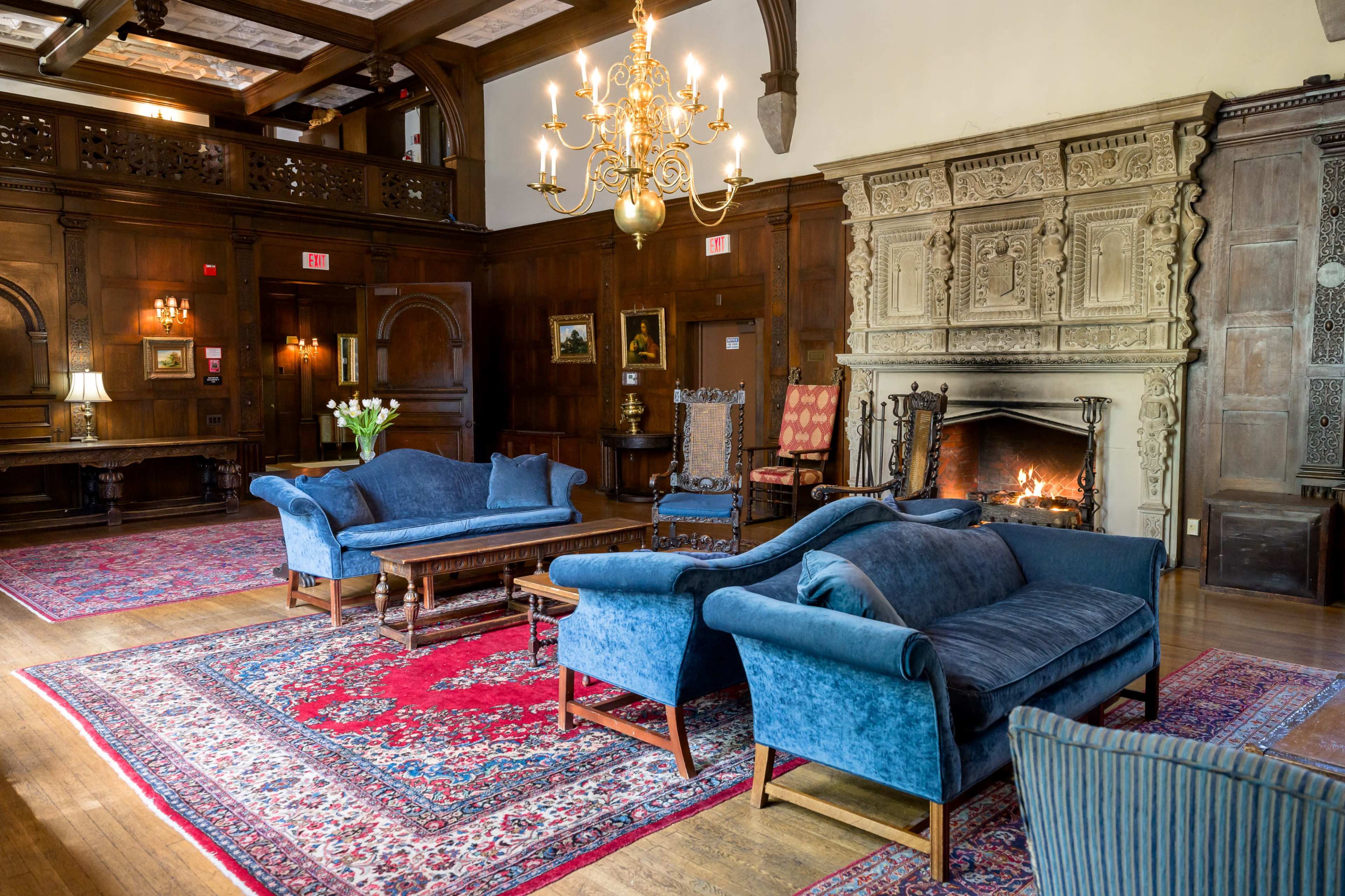 A spacious living room featuring blue fabric sofas arranged around a wooden coffee table, a large area rug, and a stone fireplace with a fire burning.