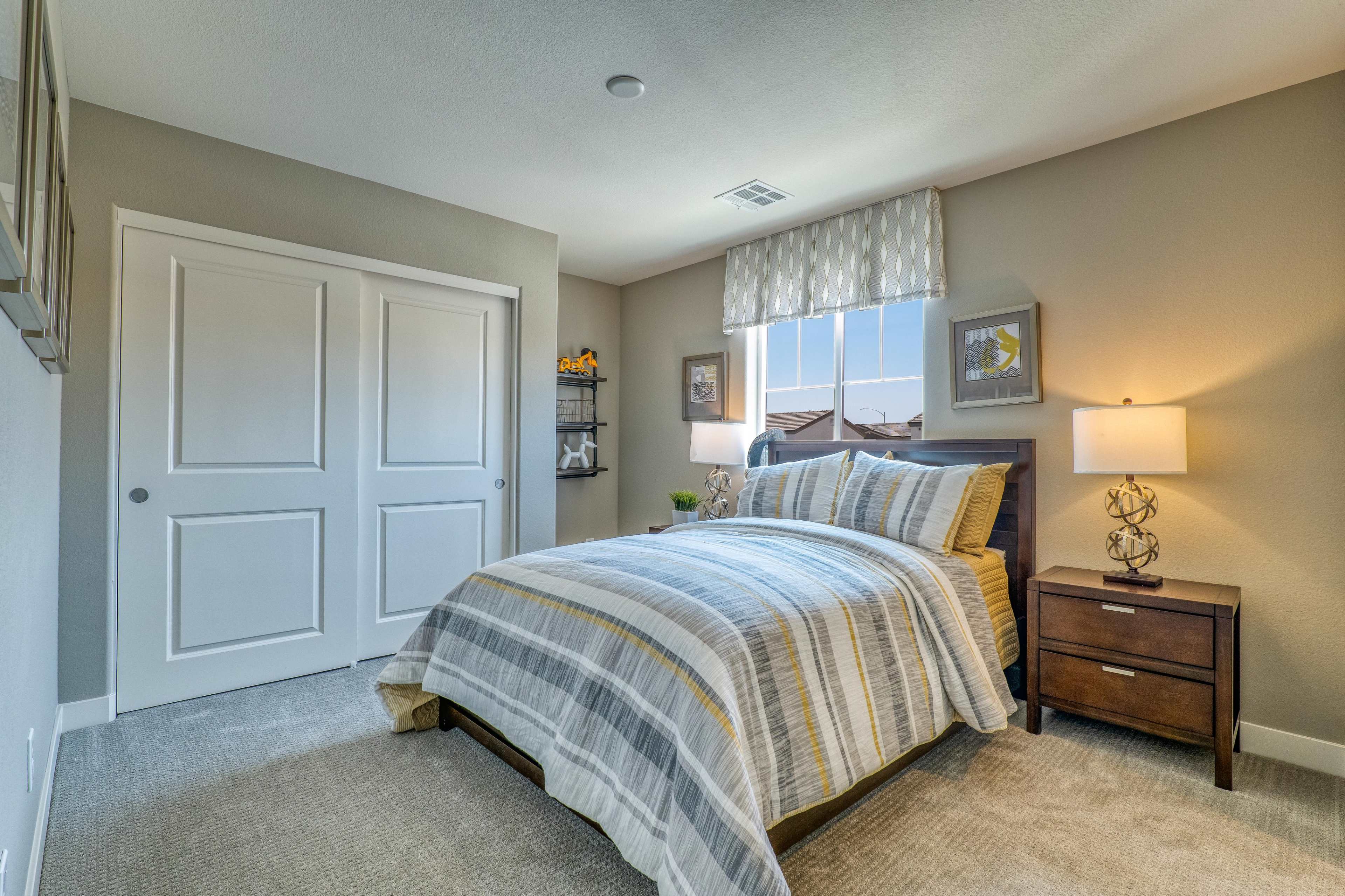 The image shows a neatly arranged bedroom featuring a bed with a striped blanket, a nightstand with a lamp, and a window with a patterned valance.