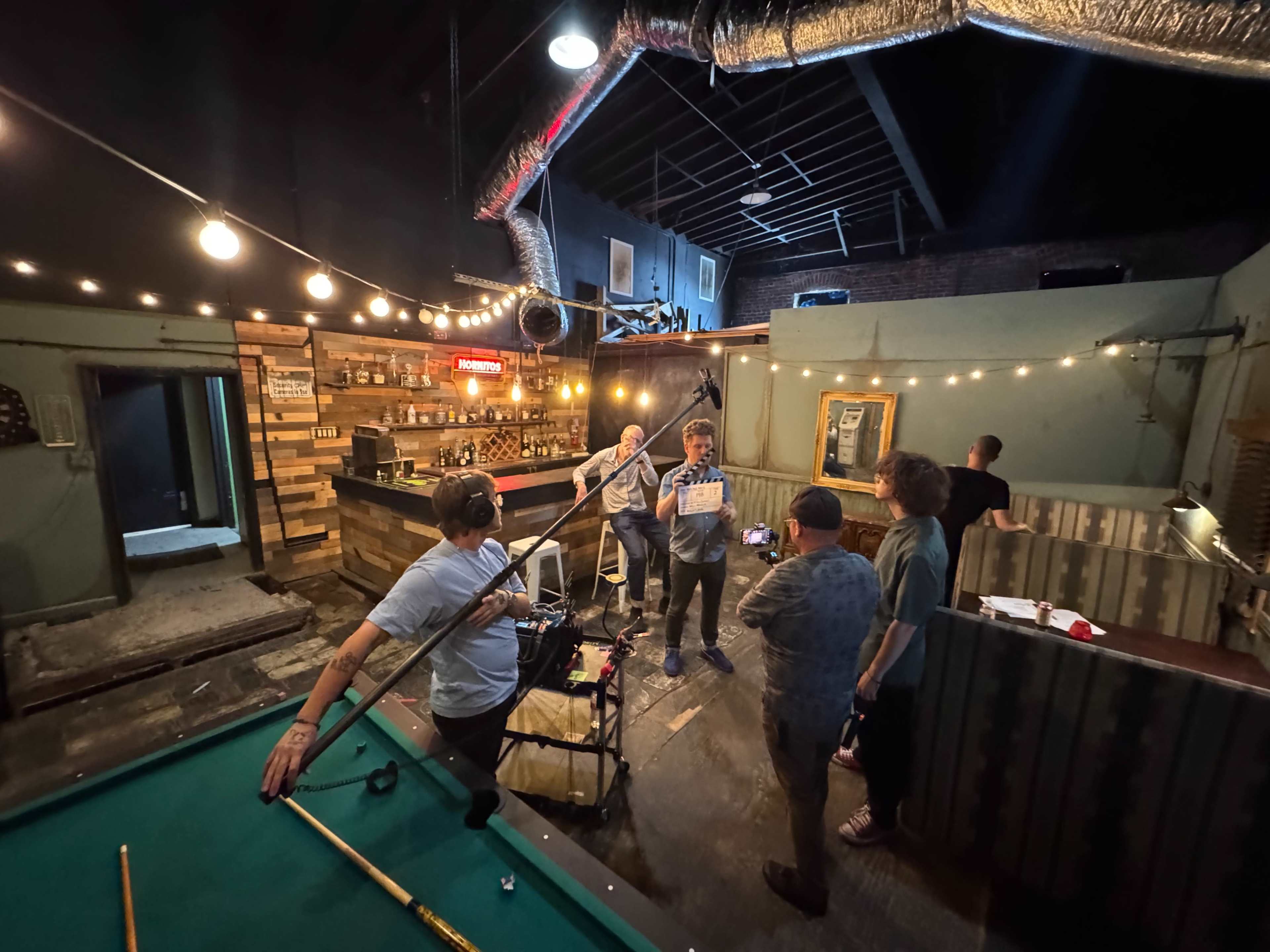 A group of individuals is gathered around a pool table in a dimly lit bar setting, engaged in conversation while another person records the scene.