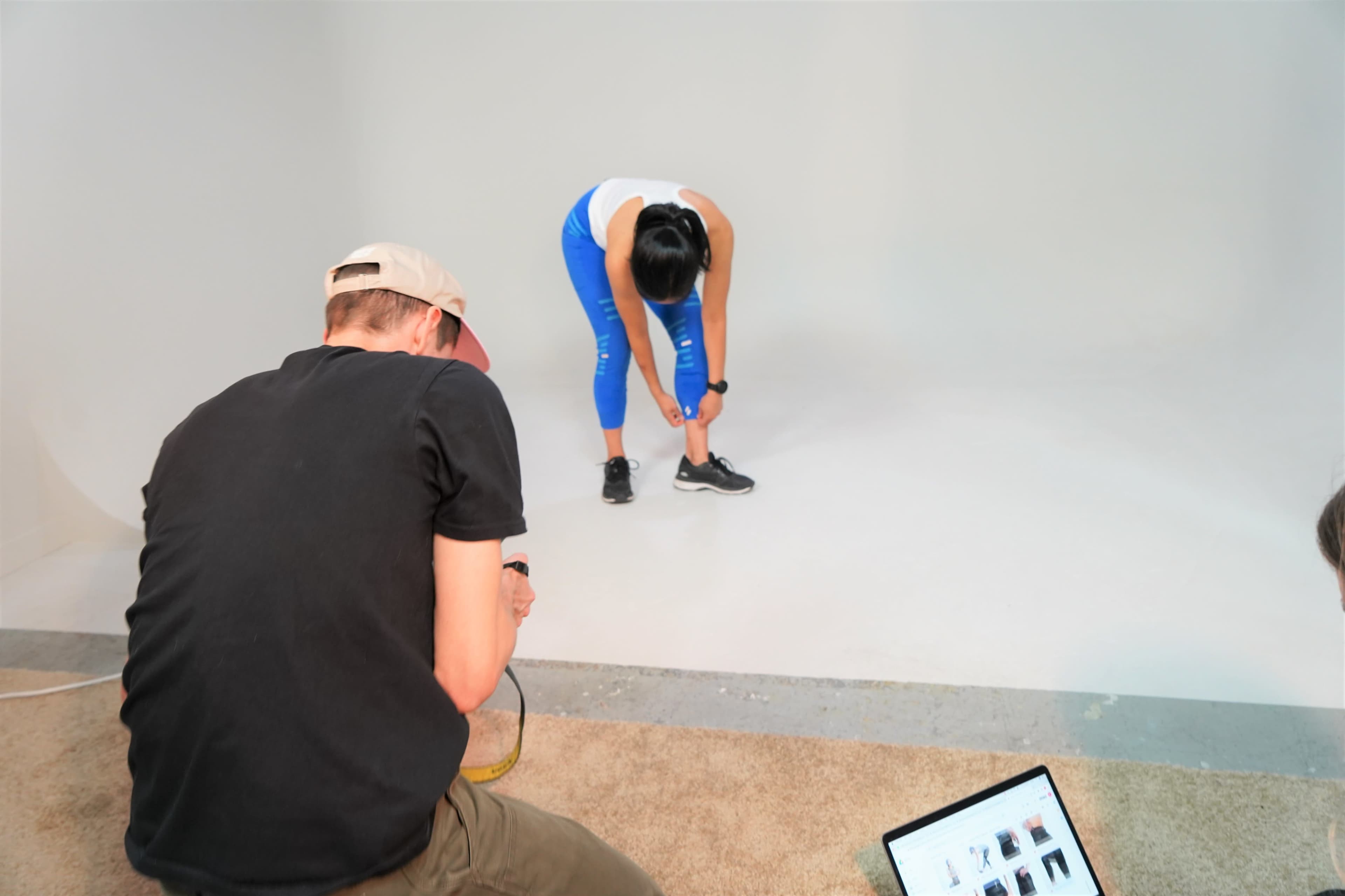 A person is bending down to tie their shoelaces while another individual is seated nearby with a laptop.