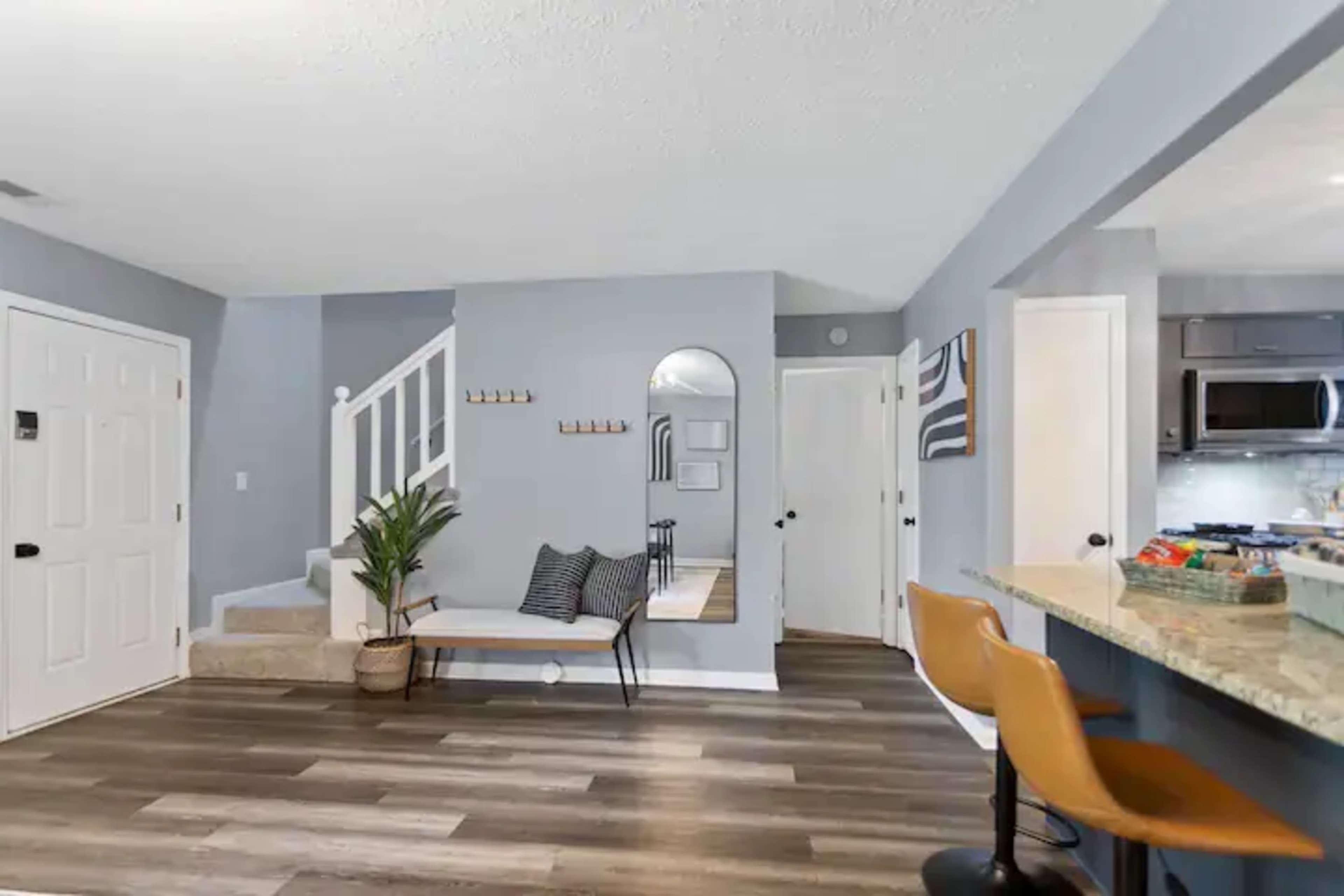 The image shows a living area with a staircase, a bench with cushions, a mirror on the wall, and bar stools at a kitchen counter.