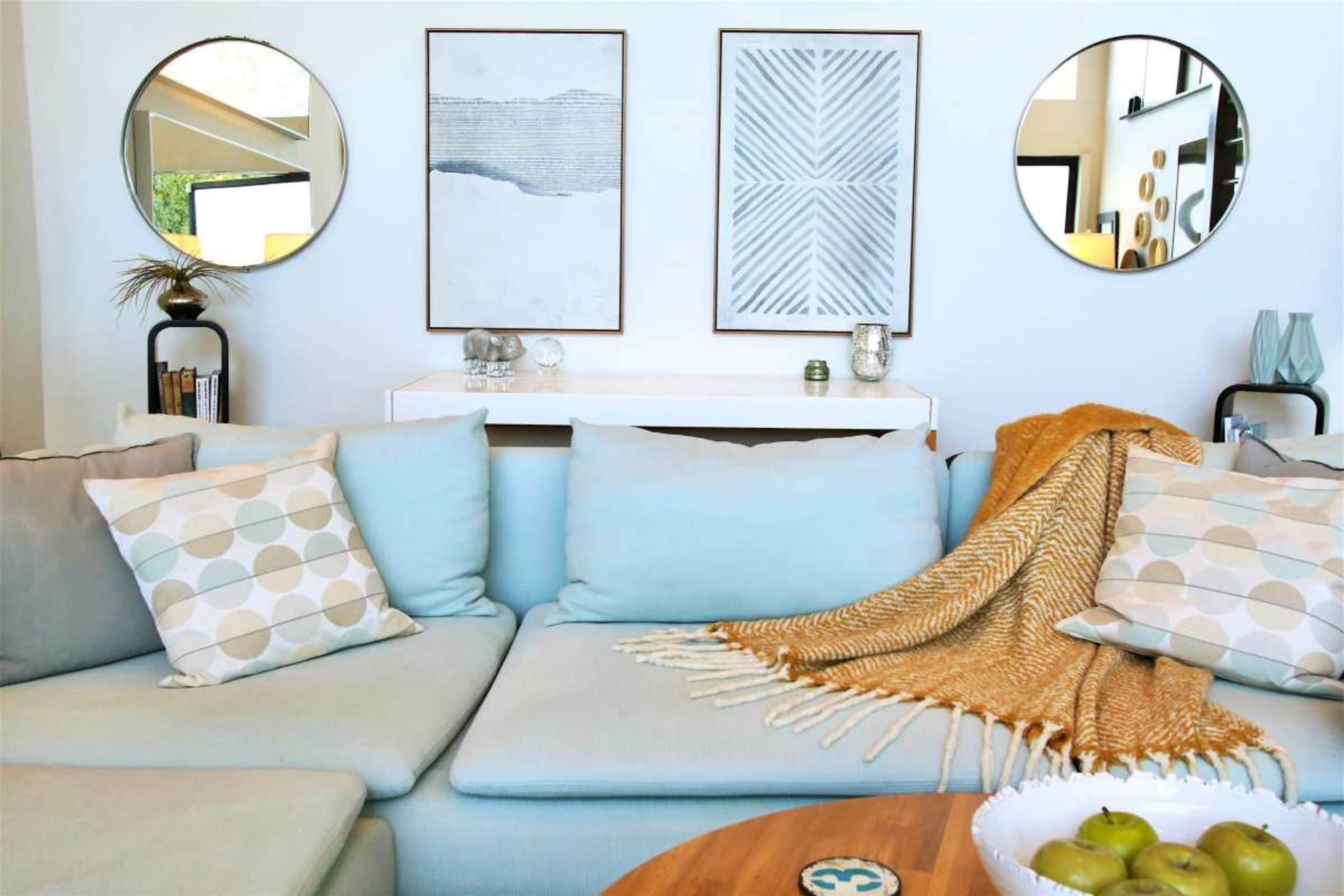 A modern living room features a light blue sectional sofa with patterned cushions, two circular mirrors on the wall, and framed artwork above a white cabinet.