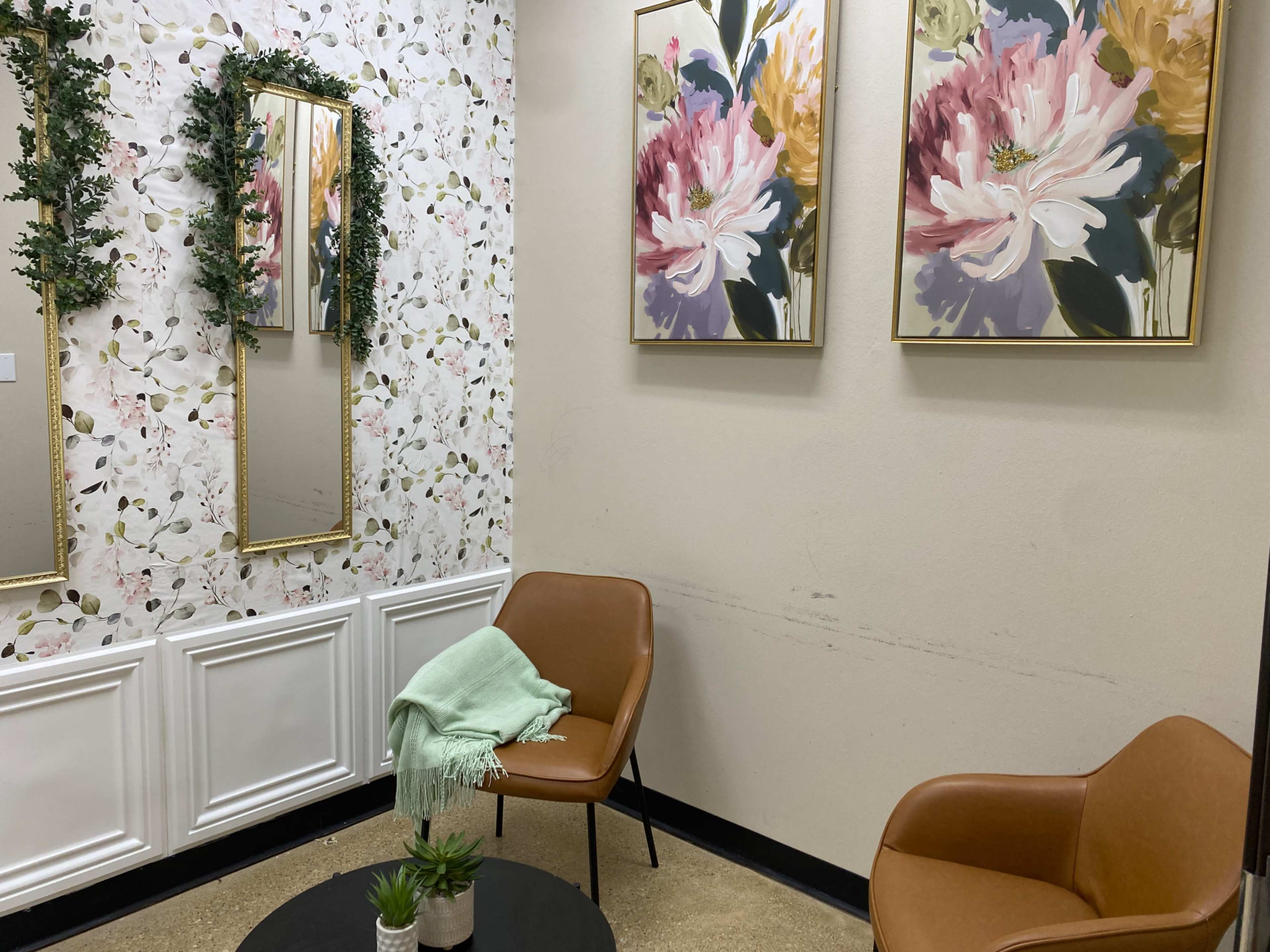 The image shows a small sitting area featuring two brown chairs, a round black coffee table, floral artwork on the walls, and mirrors adorned with foliage.